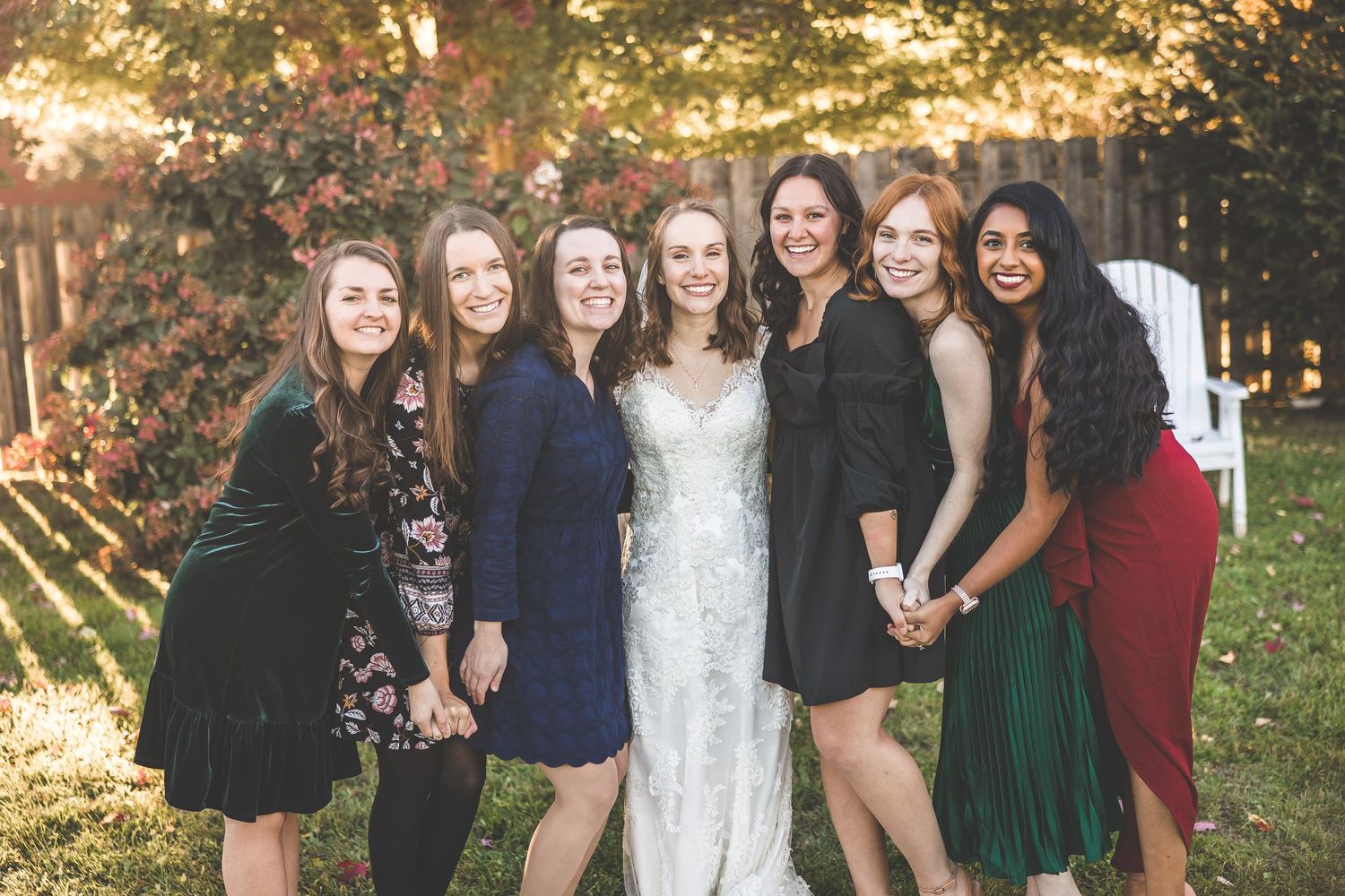 Group of women in cocktail dresses pose together at outdoor wedding celebration.