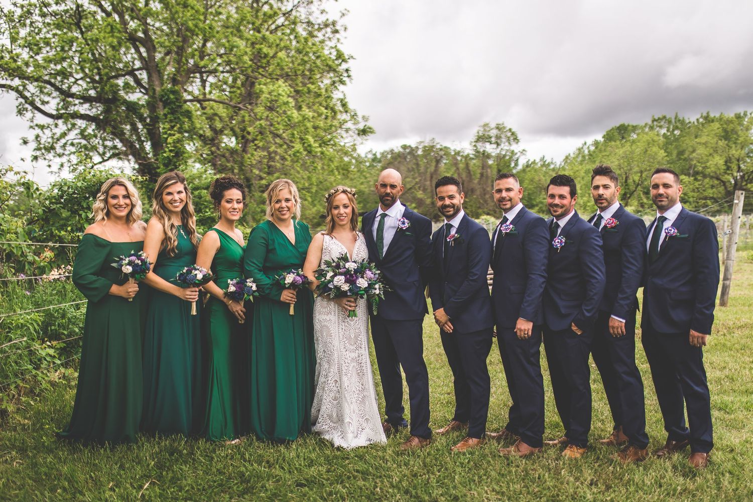 Wedding party in navy and emerald attire poses together on grassy hillside.