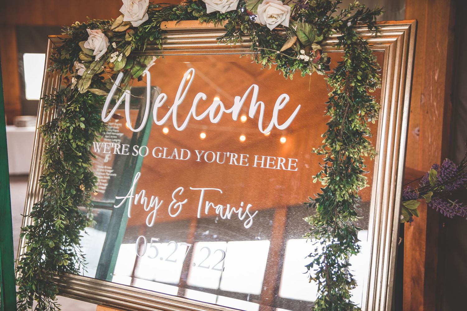Rustic welcome sign with greenery decorations hangs in wooden venue entrance.