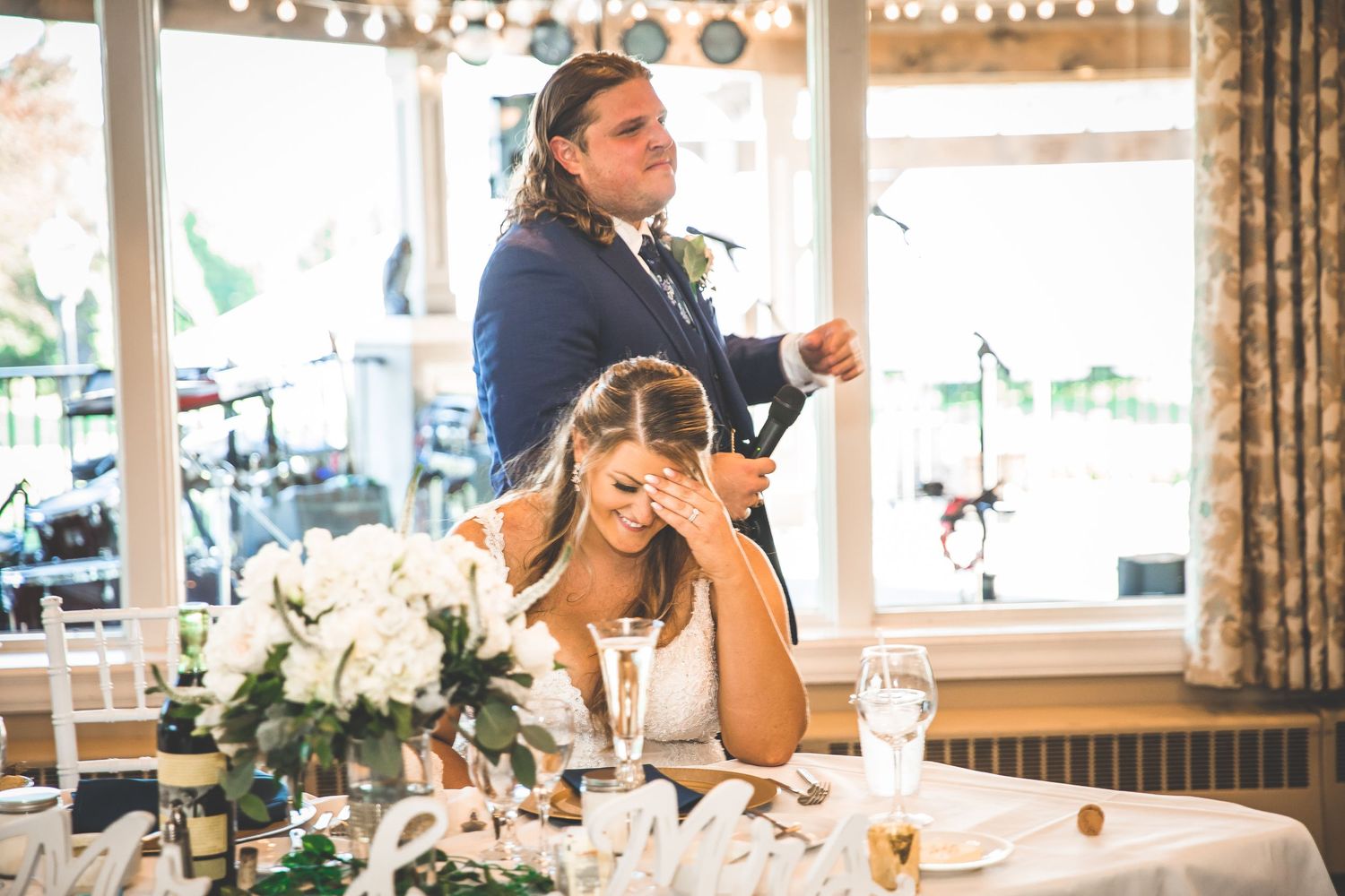 Sunlit wedding reception venue with elegant white floral centerpieces and draped windows.