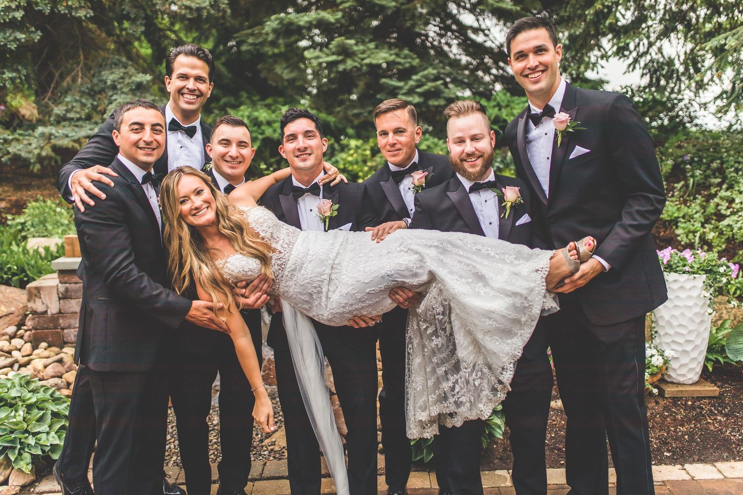 Groomsmen in black tuxedos playfully lifting bride in white lace gown outdoors.