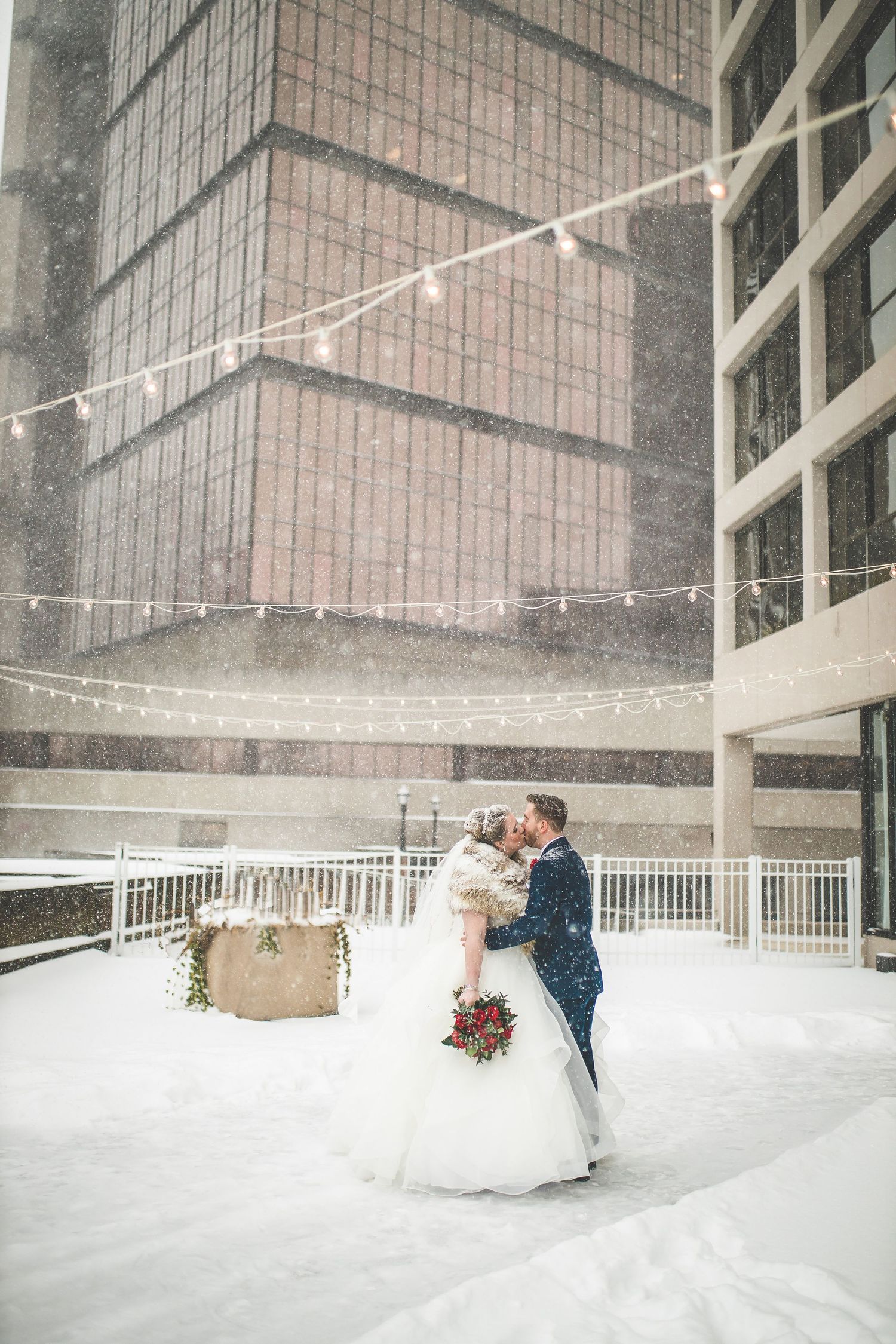 Romantic winter wedding portrait with couple embracing under string lights as snow falls.