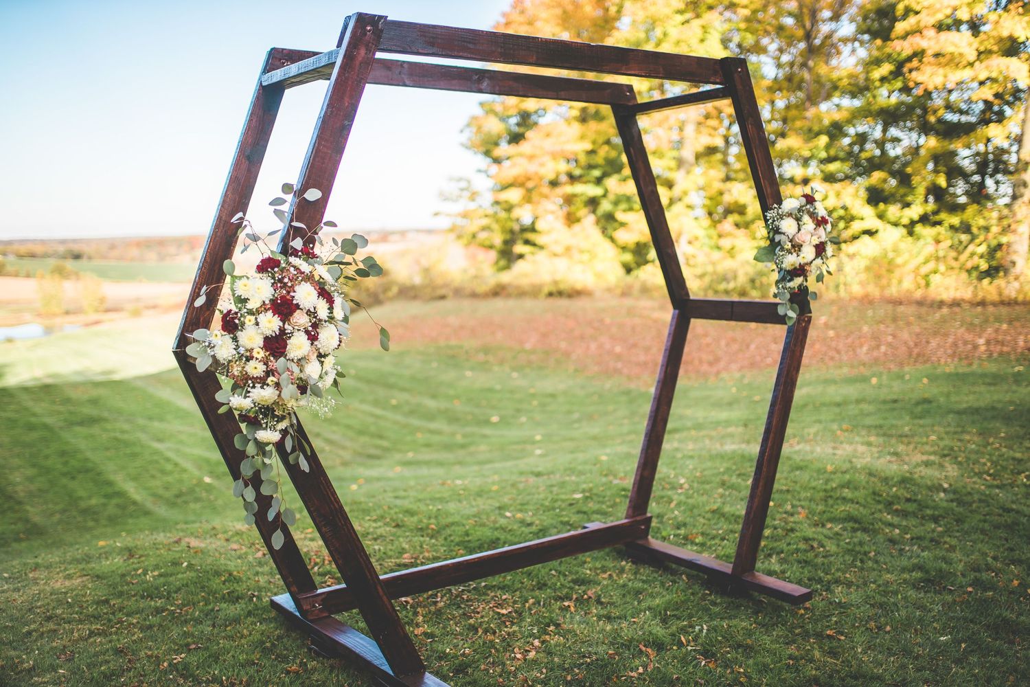 Modern hexagonal wedding arch decorated with floral arrangements against autumn landscape.