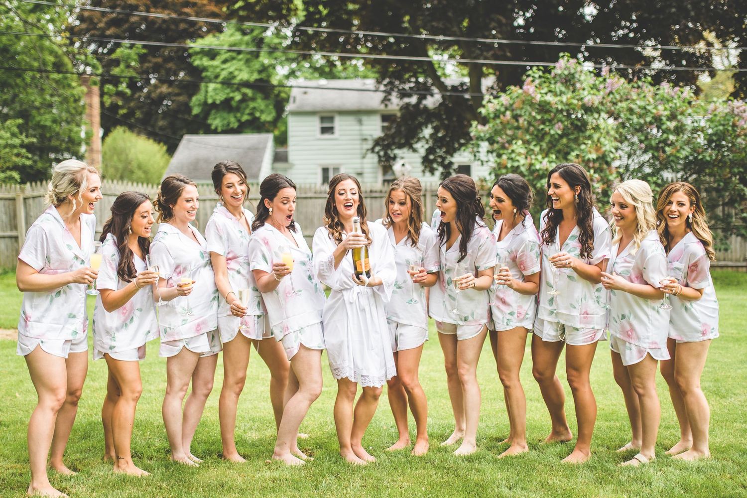 Group of bridesmaids in white robes pose together in a garden setting.