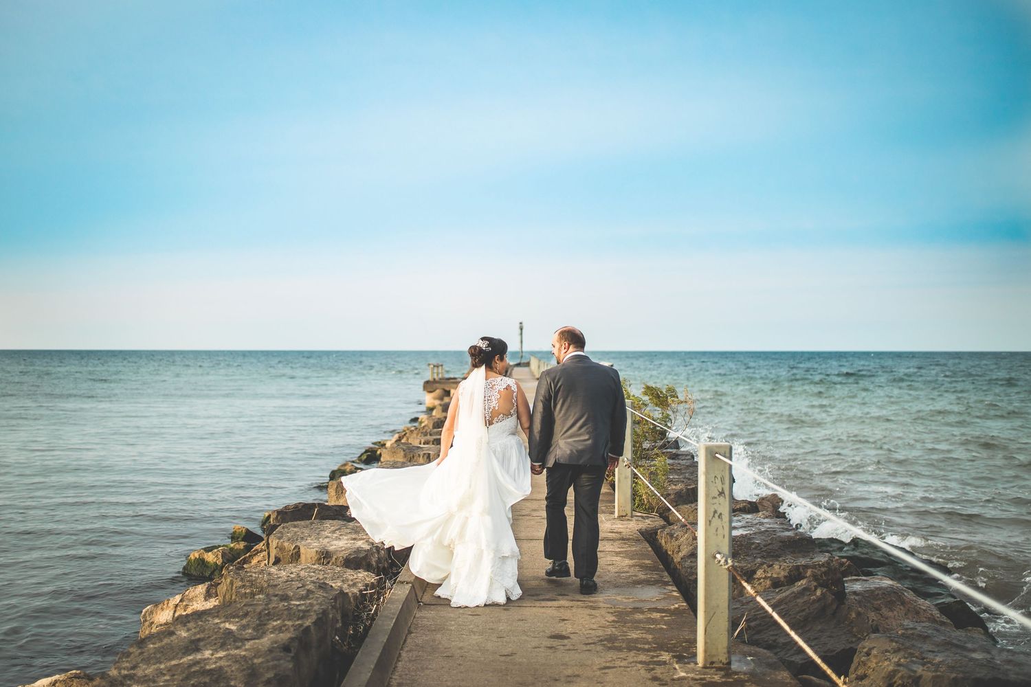 Wedding couple sits on a rocky pier overlooking the ocean at sunset.