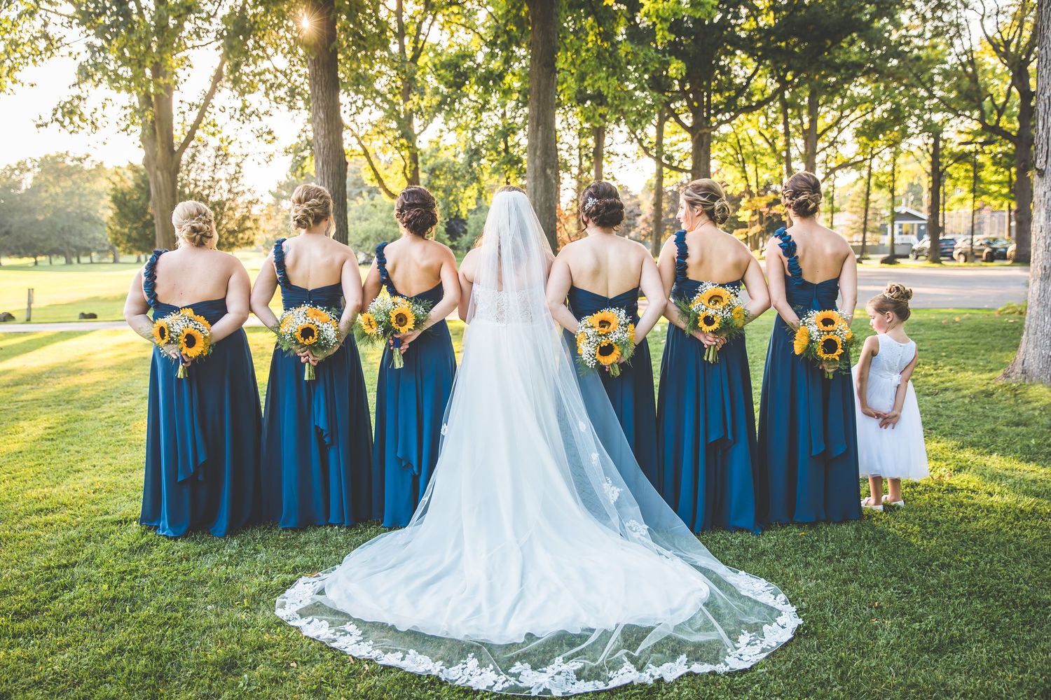 Wedding party in navy blue dresses poses together on a sunny lawn with trees in the background.