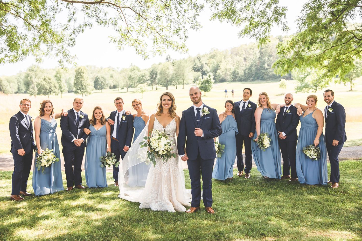 Wedding party in blue dresses and dark suits poses on a sunny lawn.