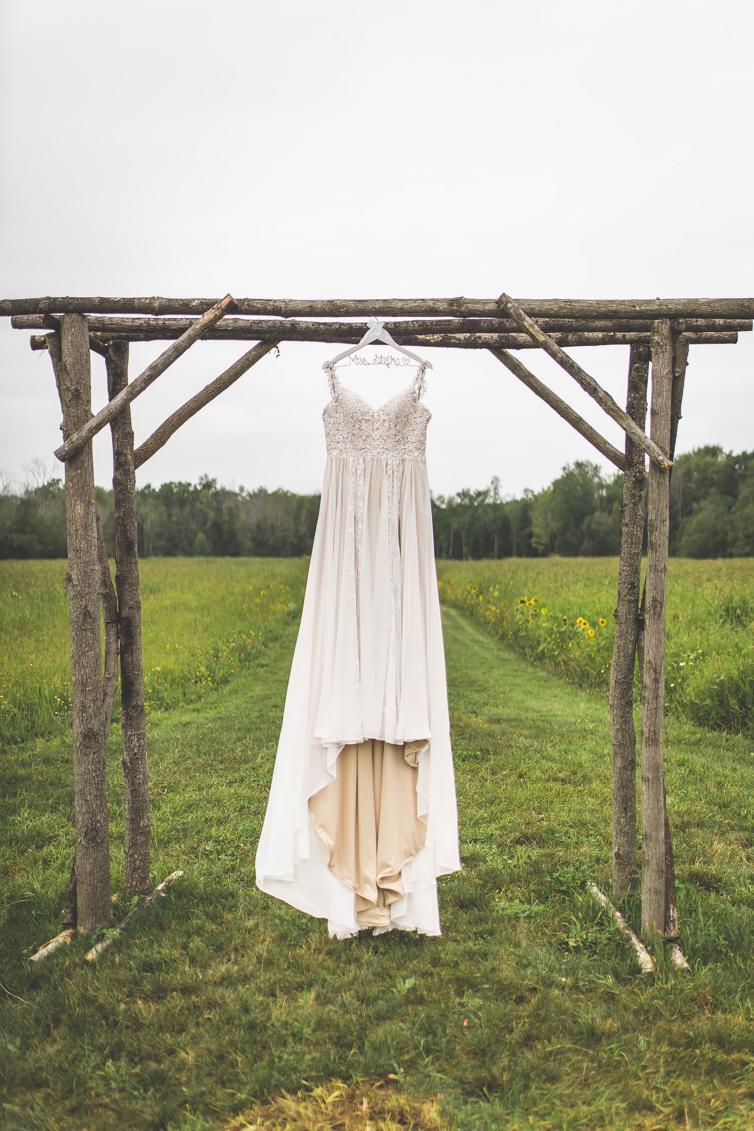 White wedding dress hanging on rustic wooden arch in green countryside field.
