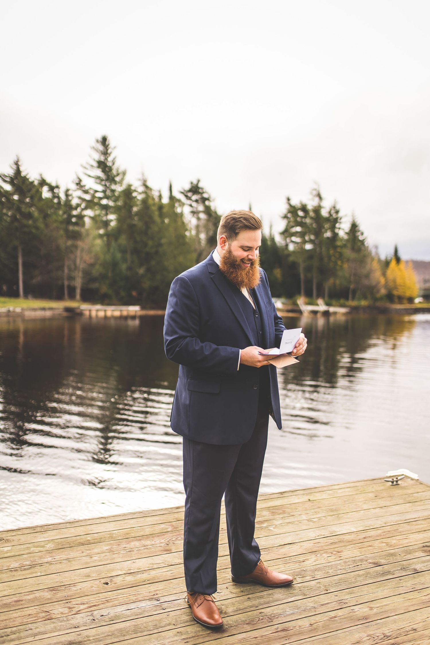 A bearded figure in a navy suit reads from paper while standing on a lakeside dock.