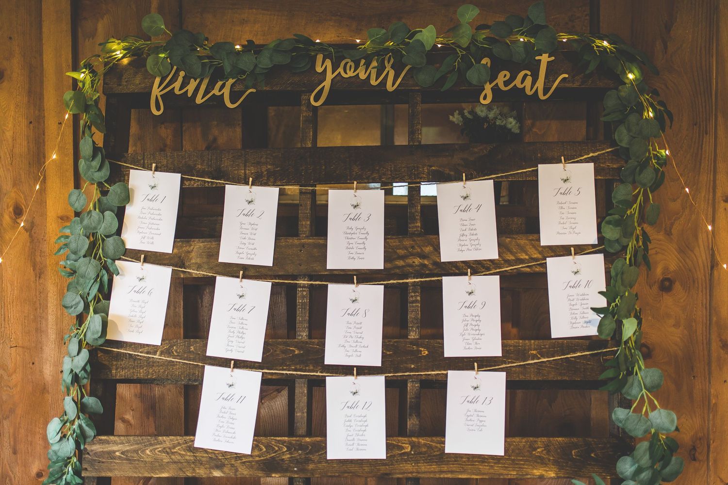 Wedding seating chart with cards hanging on twine against wooden wall with greenery.