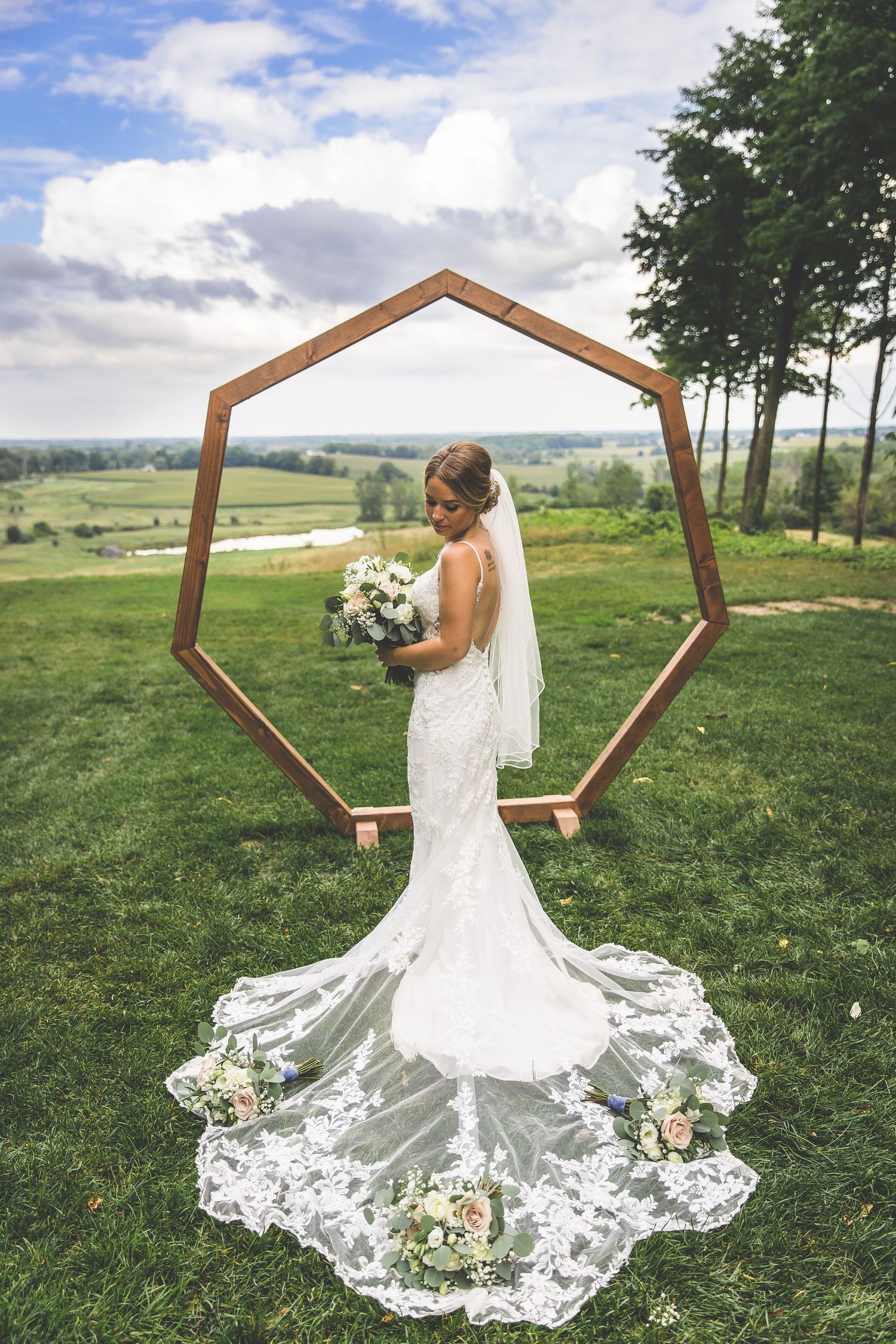 A bride in a lace wedding dress poses in front of a wooden hexagonal arch on a grassy hilltop.