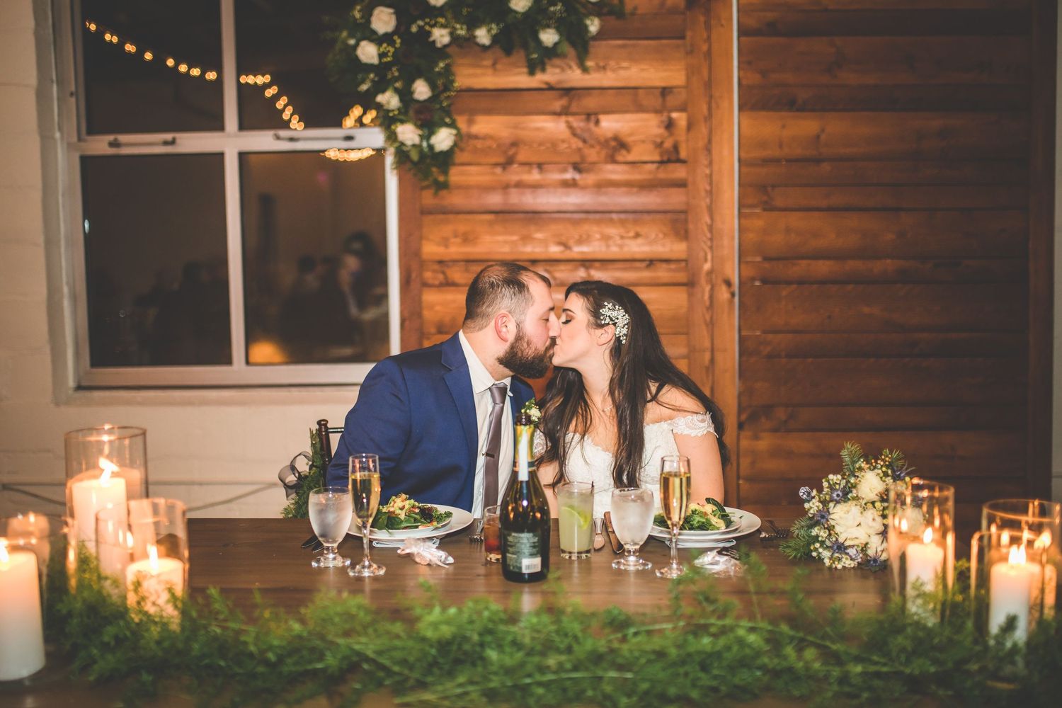 Rustic wedding reception table with candles, floral centerpieces and wooden wall backdrop.