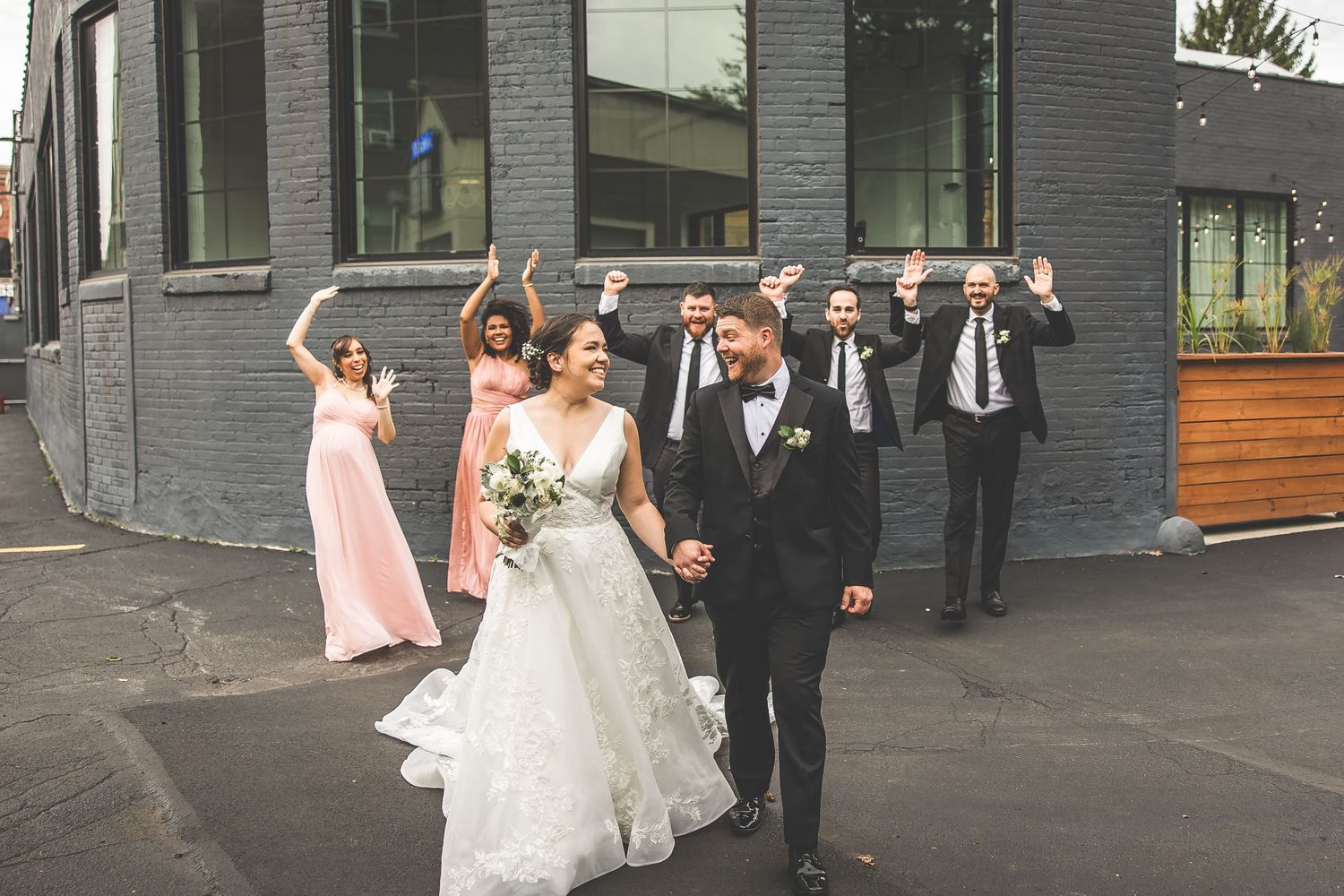 Wedding party lined up outdoors against modern building with groomsmen in dark suits.