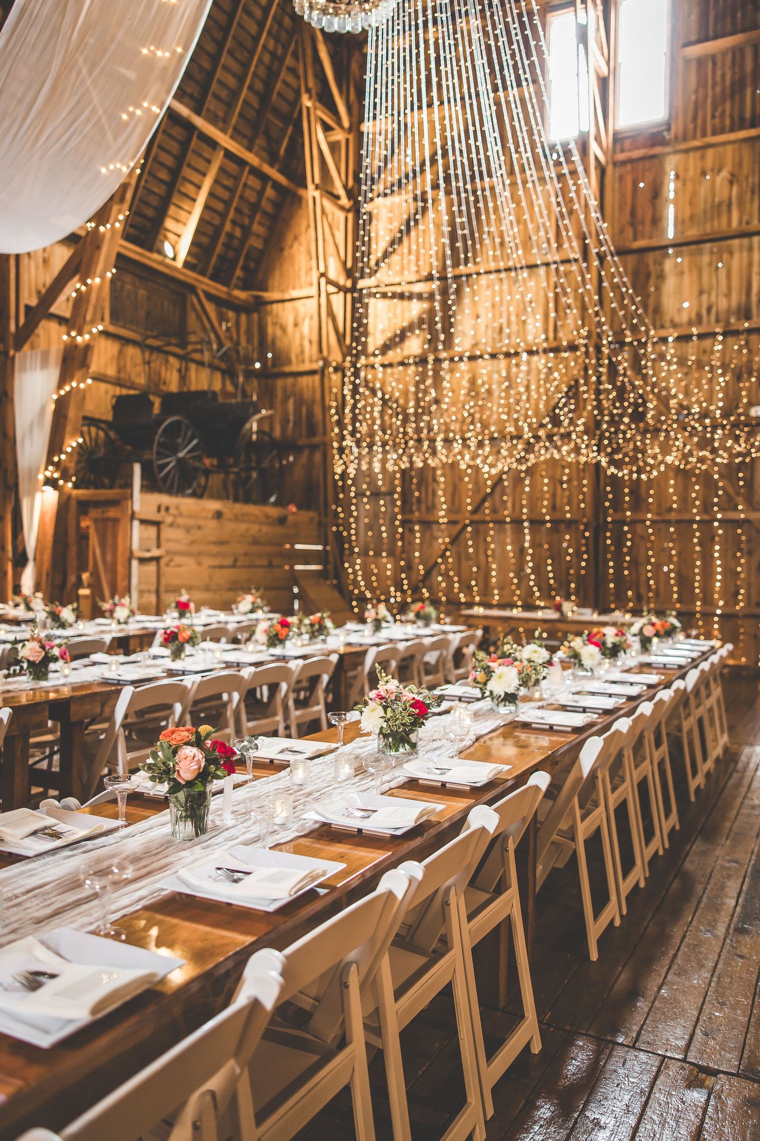 Barn wedding reception setup with twinkling lights, white draping, and long wooden tables.