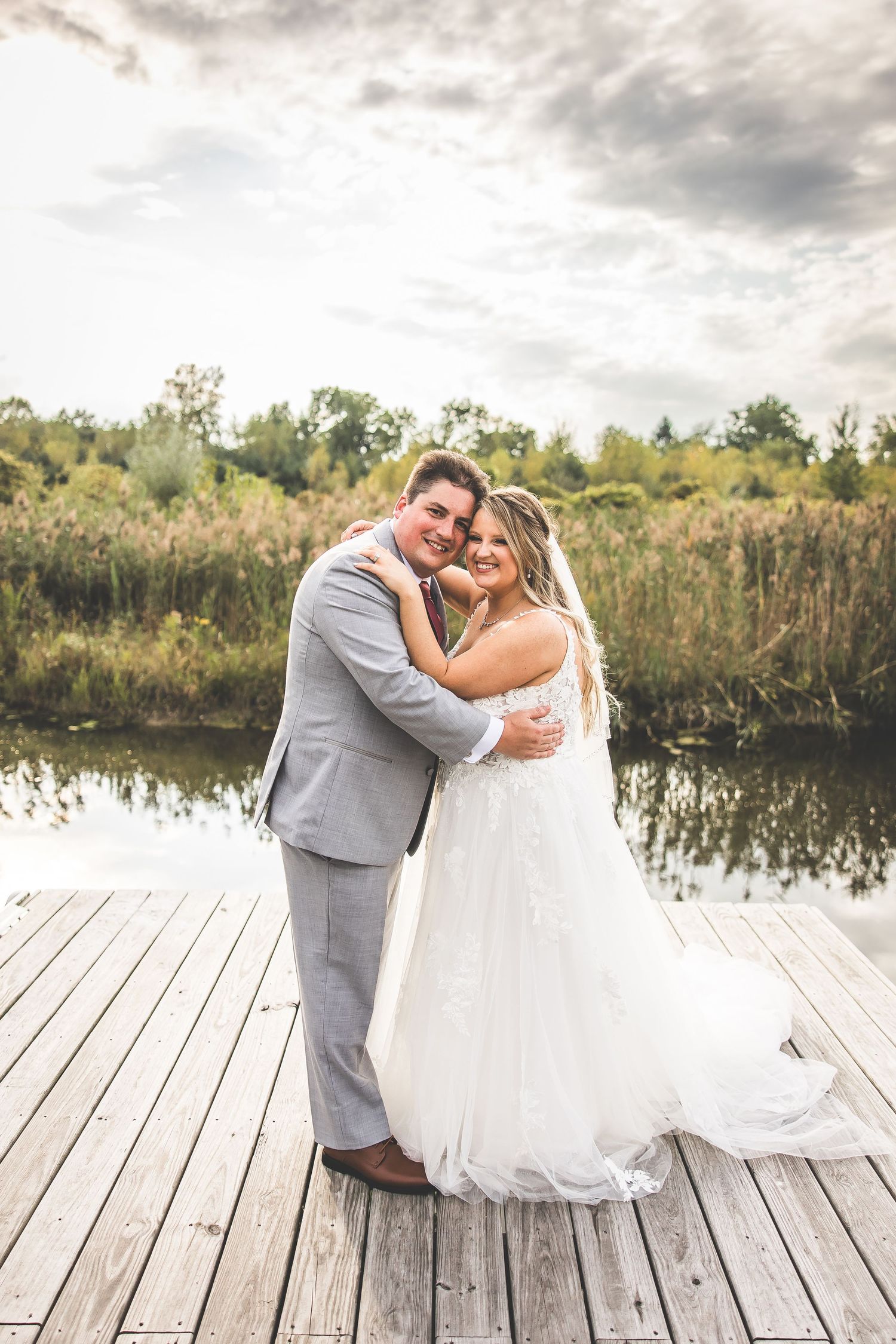 A romantic wedding couple poses together on a wooden dock by a tranquil pond on their special day.