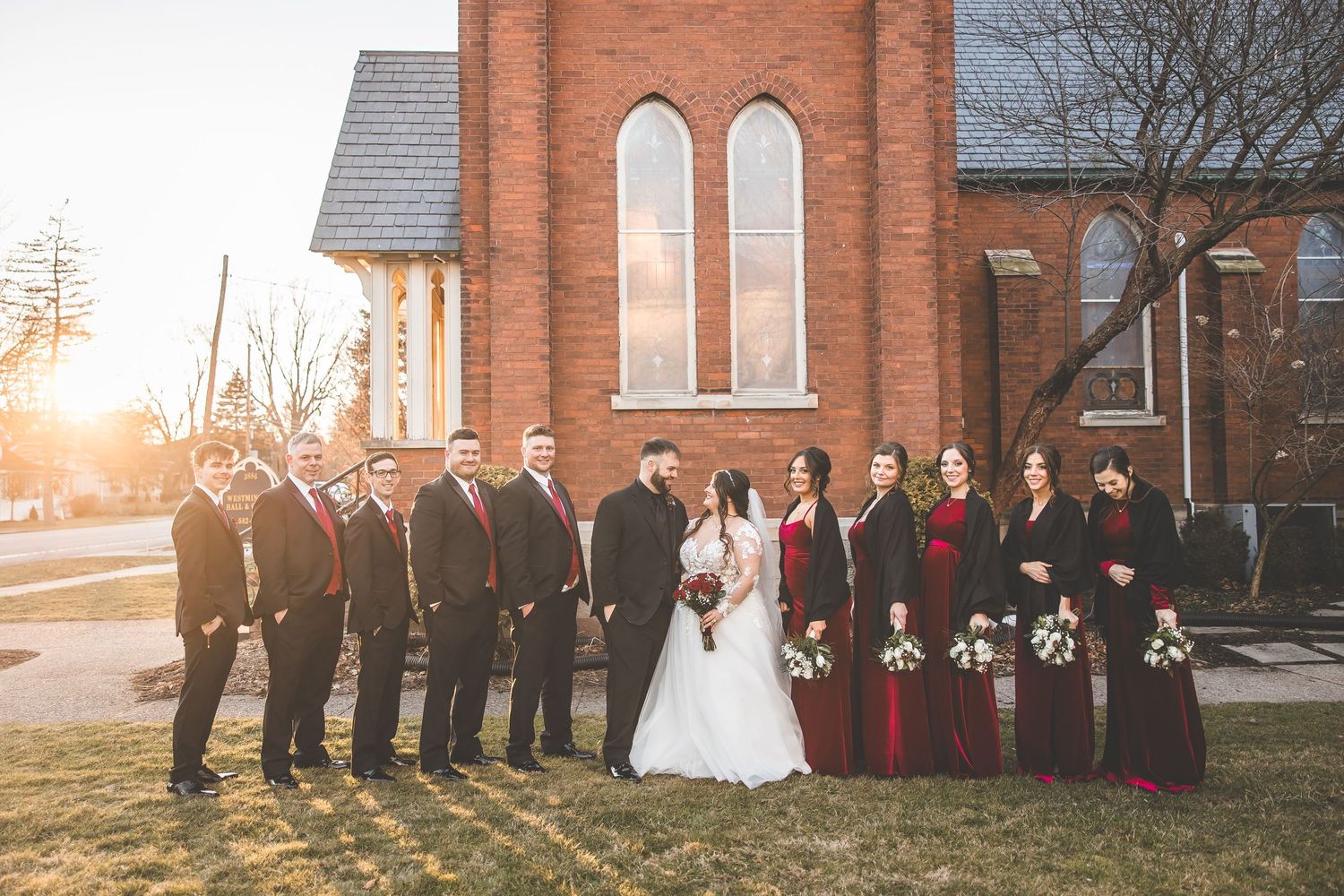 Wedding group photo with bridal party in red dresses against brick church exterior.