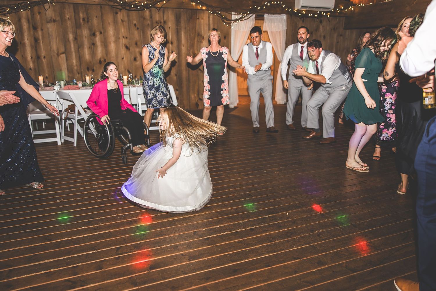 Wedding guests form a circle on a wooden dance floor with colorful lighting effects.