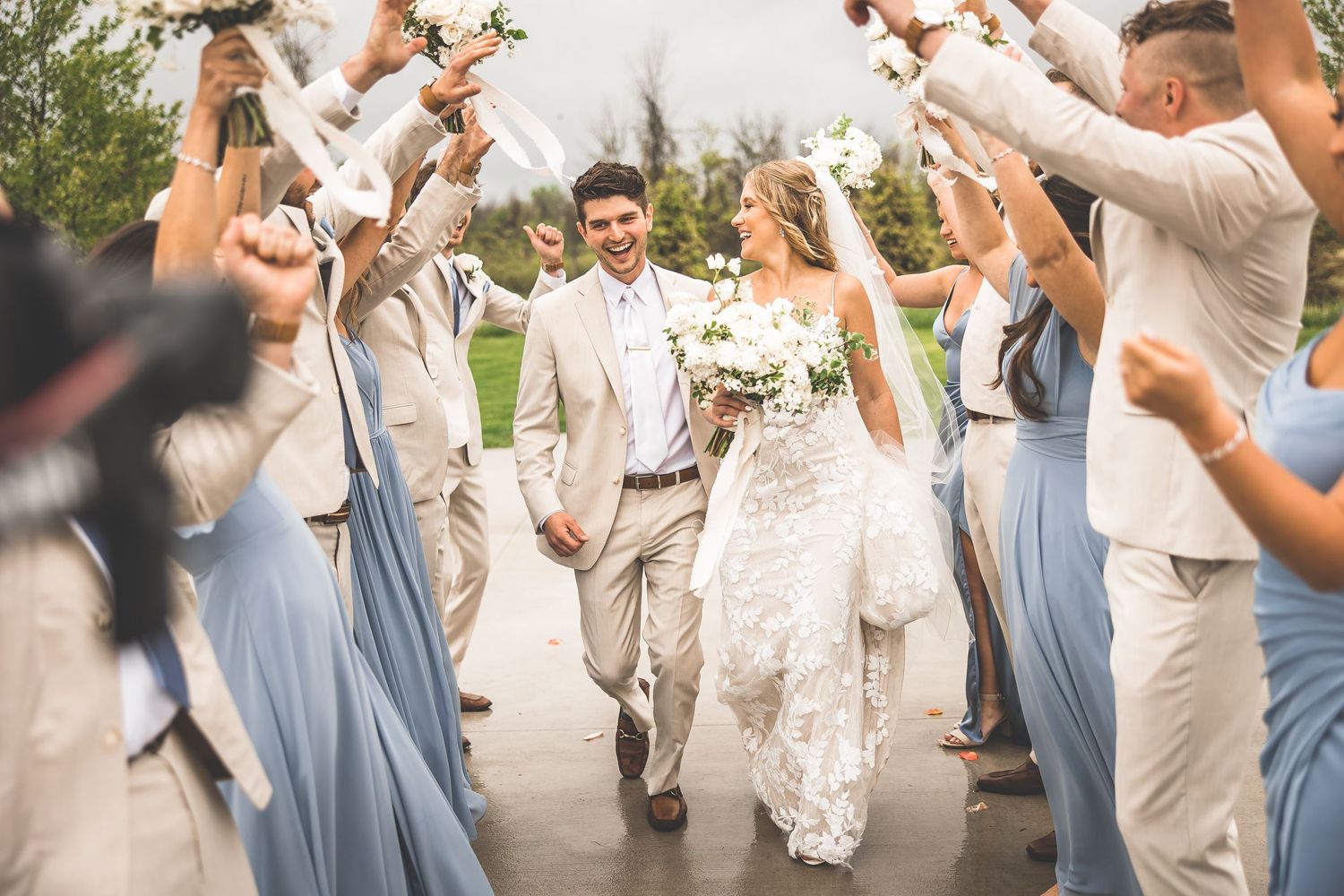 Wedding party forms celebratory tunnel with raised arms on wet concrete patio.