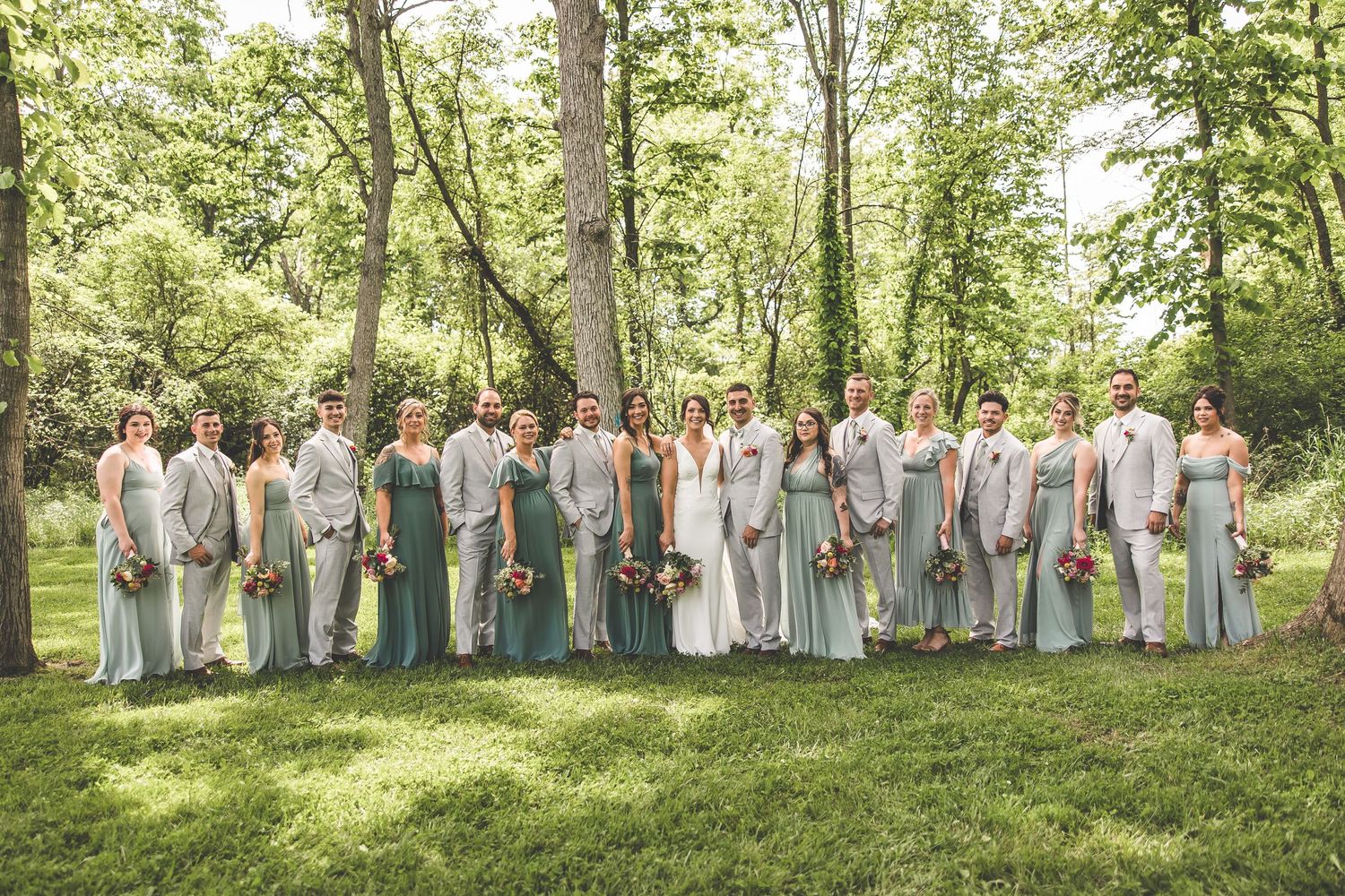 Wedding party in gray suits and sage green dresses poses on a sunny lawn.