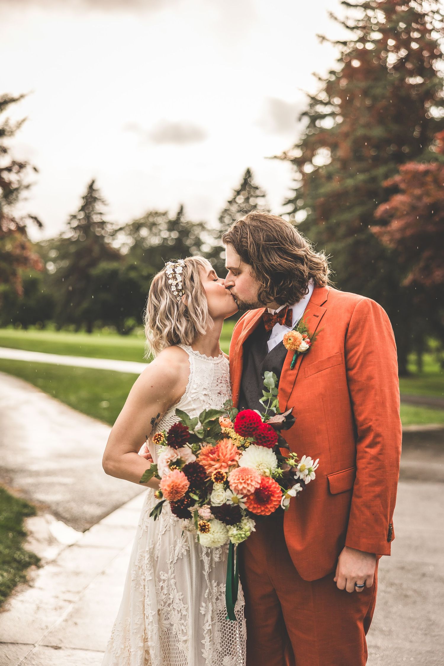 Wedding couple holding vibrant bouquets with red and peach flowers against autumn trees.
