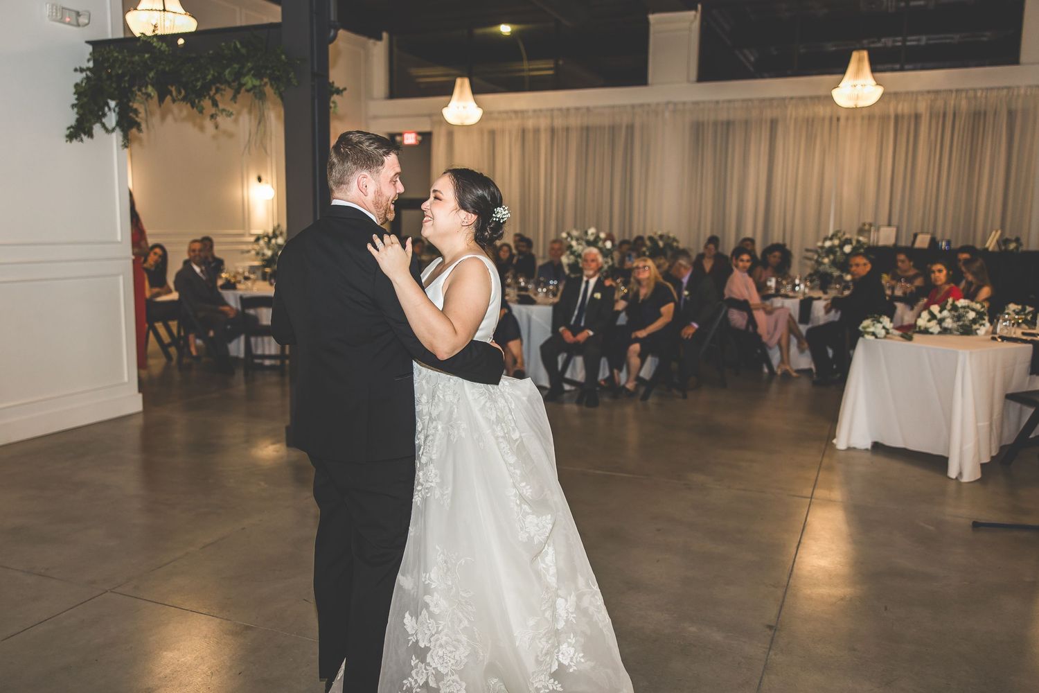 A couple dances in the spotlight while wedding guests watch from decorated tables.