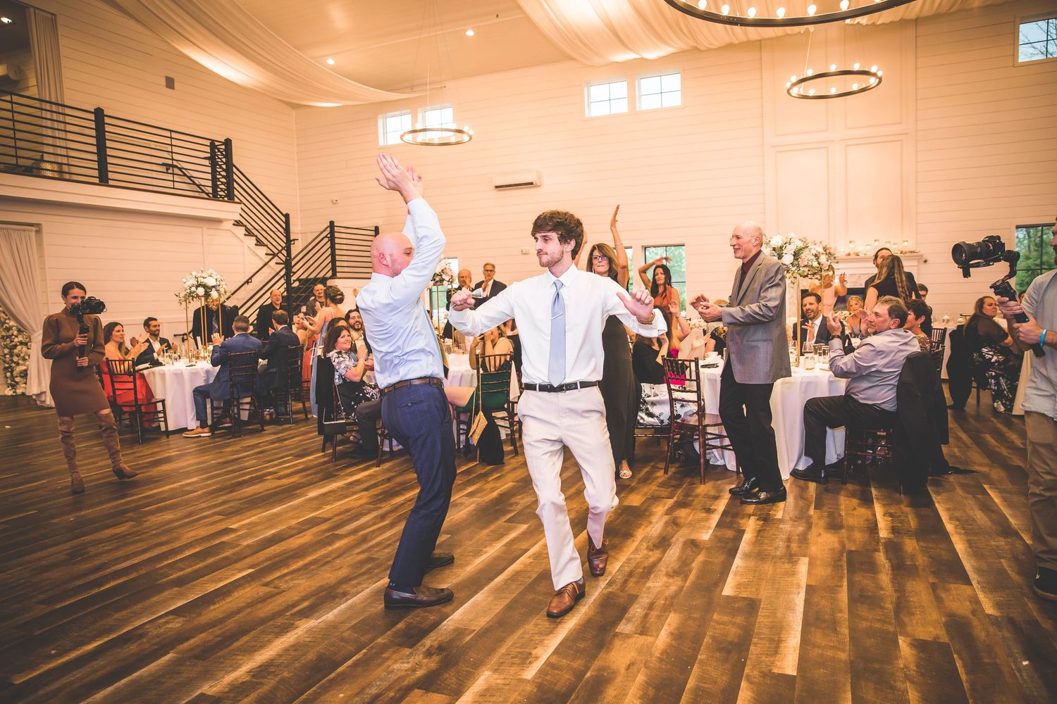 Wedding guests dancing and celebrating on wooden dance floor at indoor reception venue.