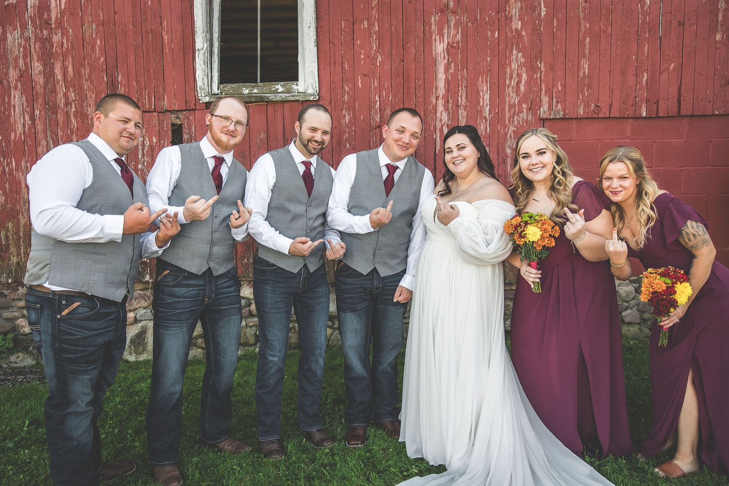 Wedding party lines up against weathered red barn wall for group photos.