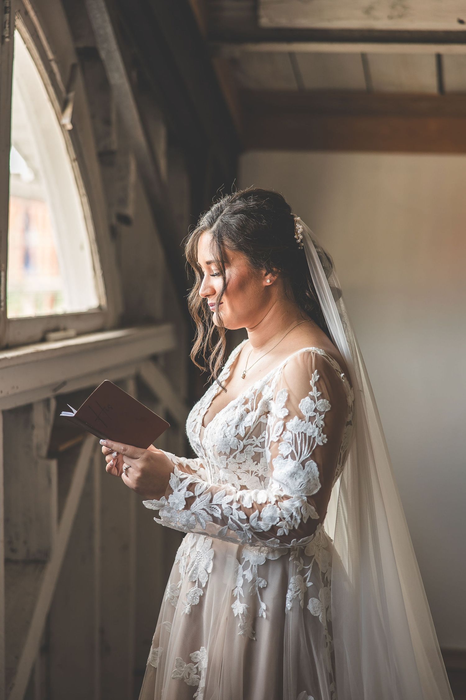 A bride in a delicate lace wedding gown with flowing veil stands in soft window light.