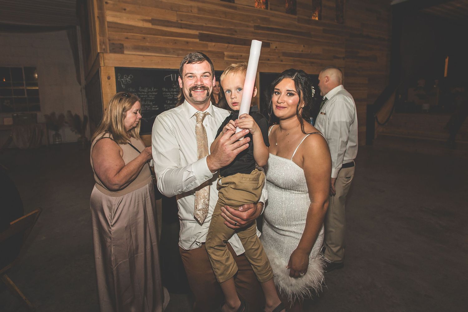 Wedding guests celebrate at a rustic barn reception with string lights and wooden walls.