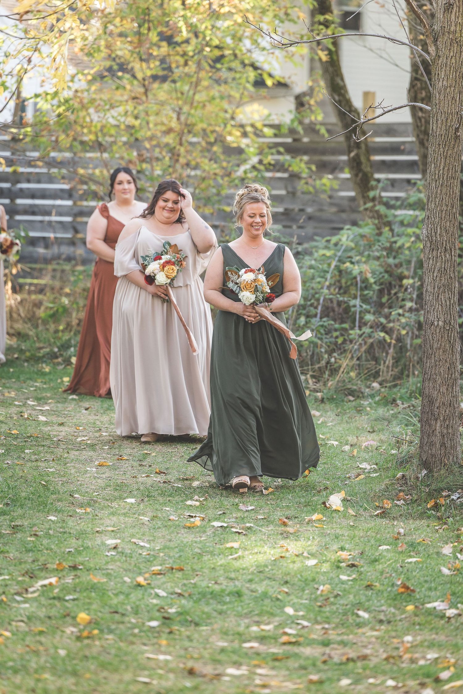 Bridesmaids in earth-toned dresses walk down outdoor aisle carrying bouquets.