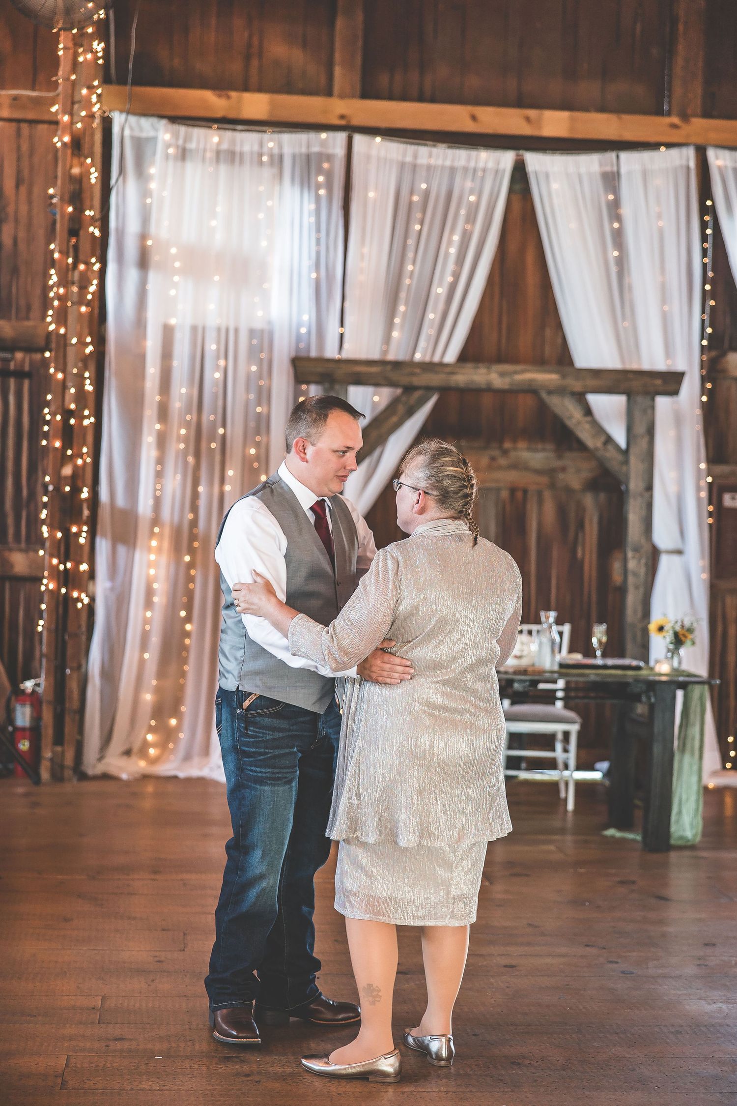 Wedding guests dancing in a beautifully decorated barn with string lights and white drapery.