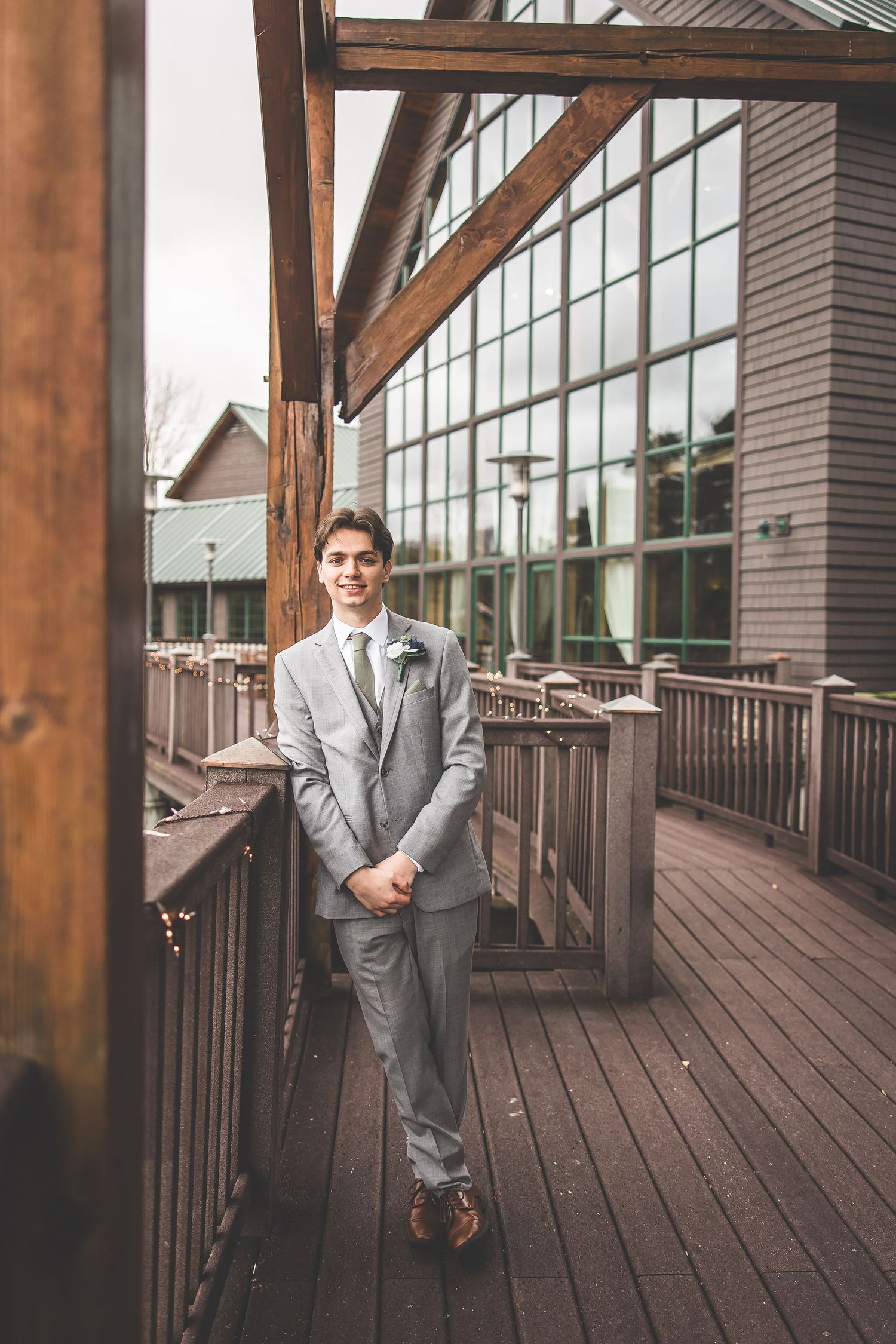 Groom in light grey suit poses on wooden deck with modern architecture.