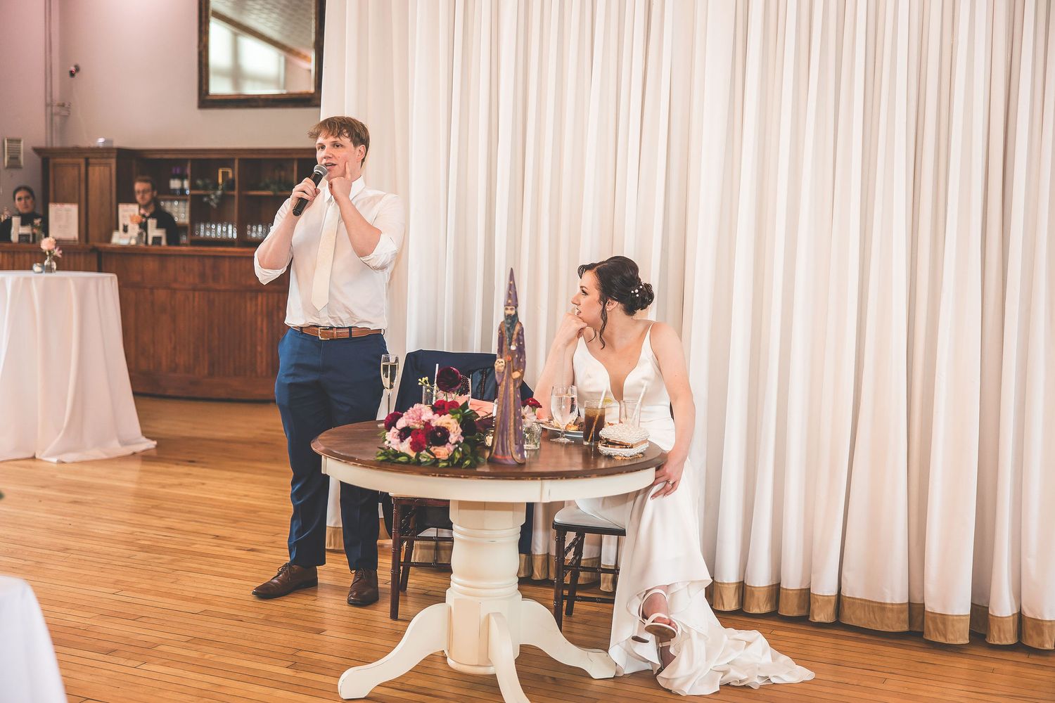 Wedding guests enjoy a celebration in an elegant venue with white drapes and wooden floors.