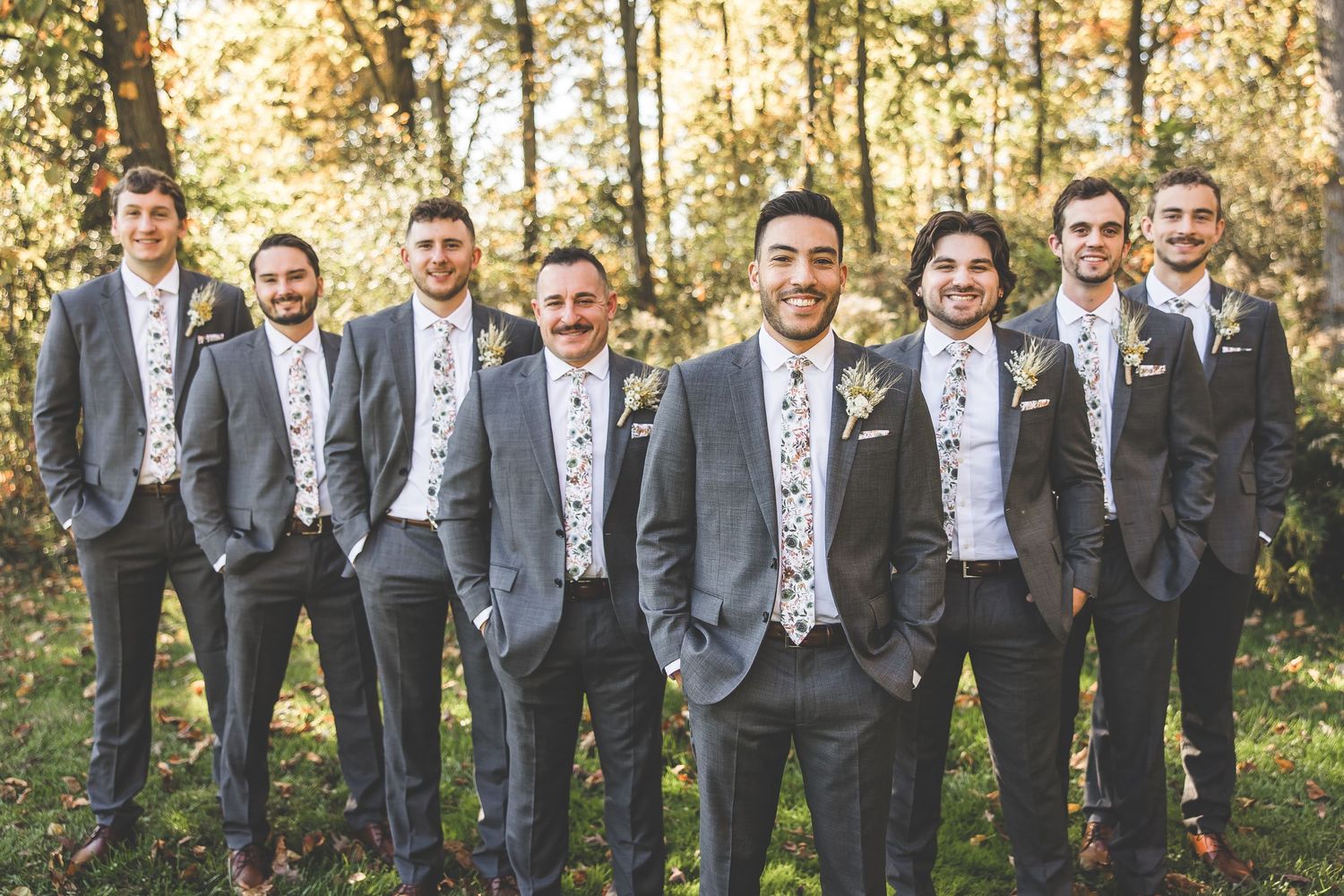 Wedding party in gray suits standing casually in a wooded area with fall foliage.