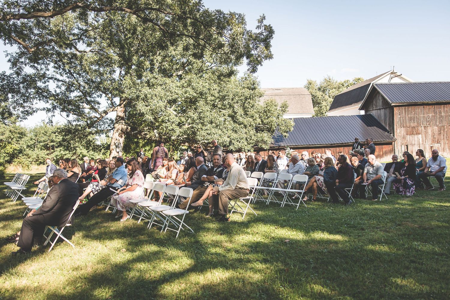 Wide view of wedding guests seated for an outdoor ceremony near a rustic barn.