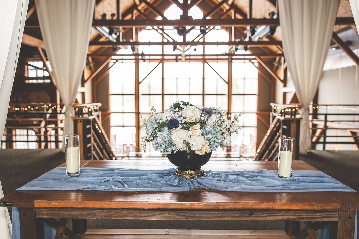 Rustic wedding table centerpiece with white floral arrangement and blue runner in barn venue.