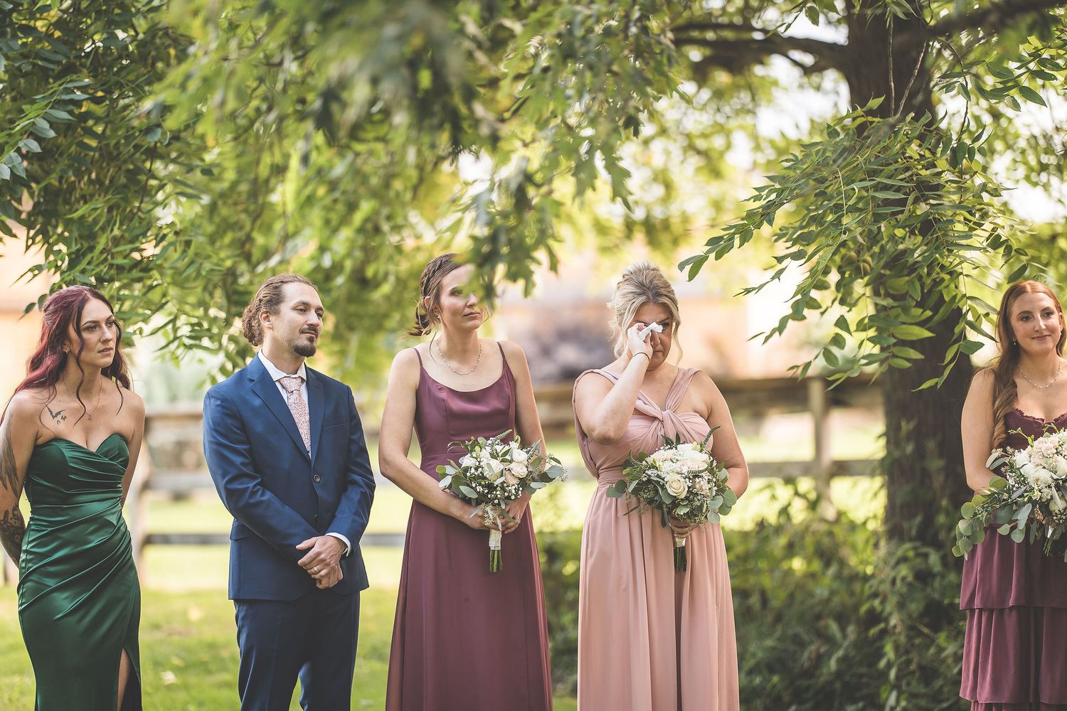 Wedding party members stand together under trees wearing pink and burgundy dresses.