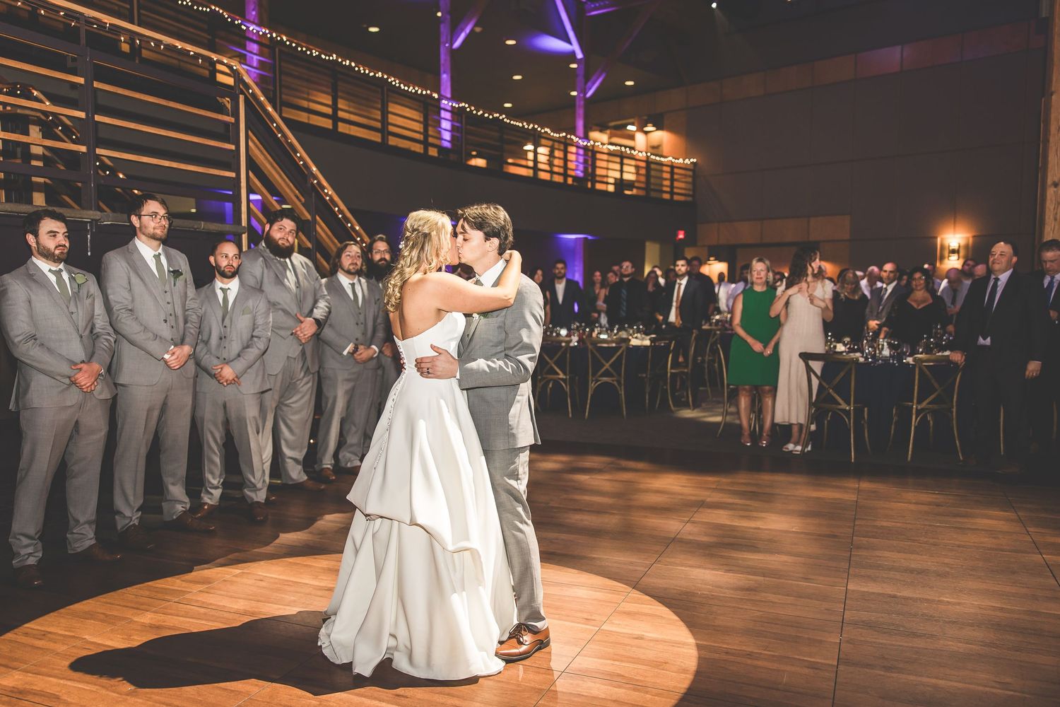 Newlyweds share their first dance surrounded by wedding party members in grey suits on an illuminated dance floor.
