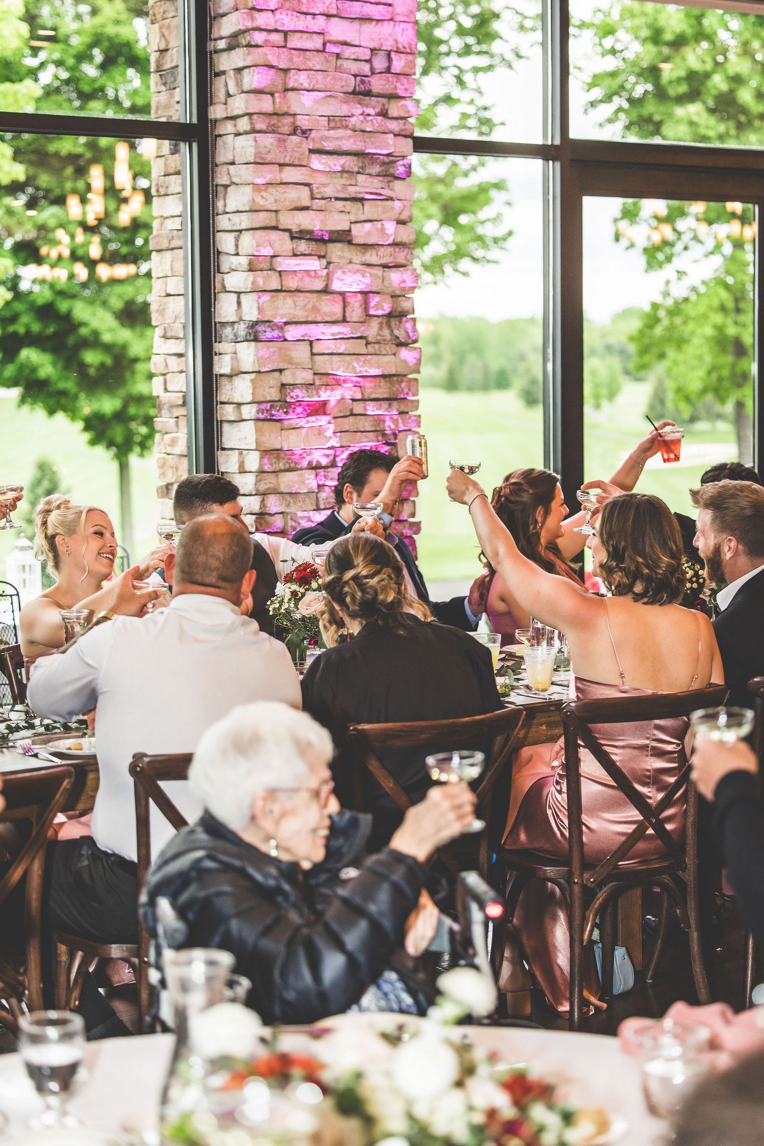 Reception guests celebrate with raised glasses inside a rustic venue with stone pillars and large windows.