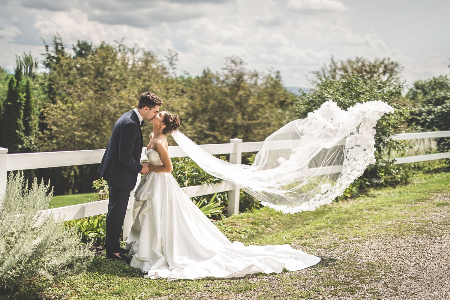 Bride and groom share a romantic kiss outdoors, with the bride's veil flowing in the wind against a lush green backdrop.