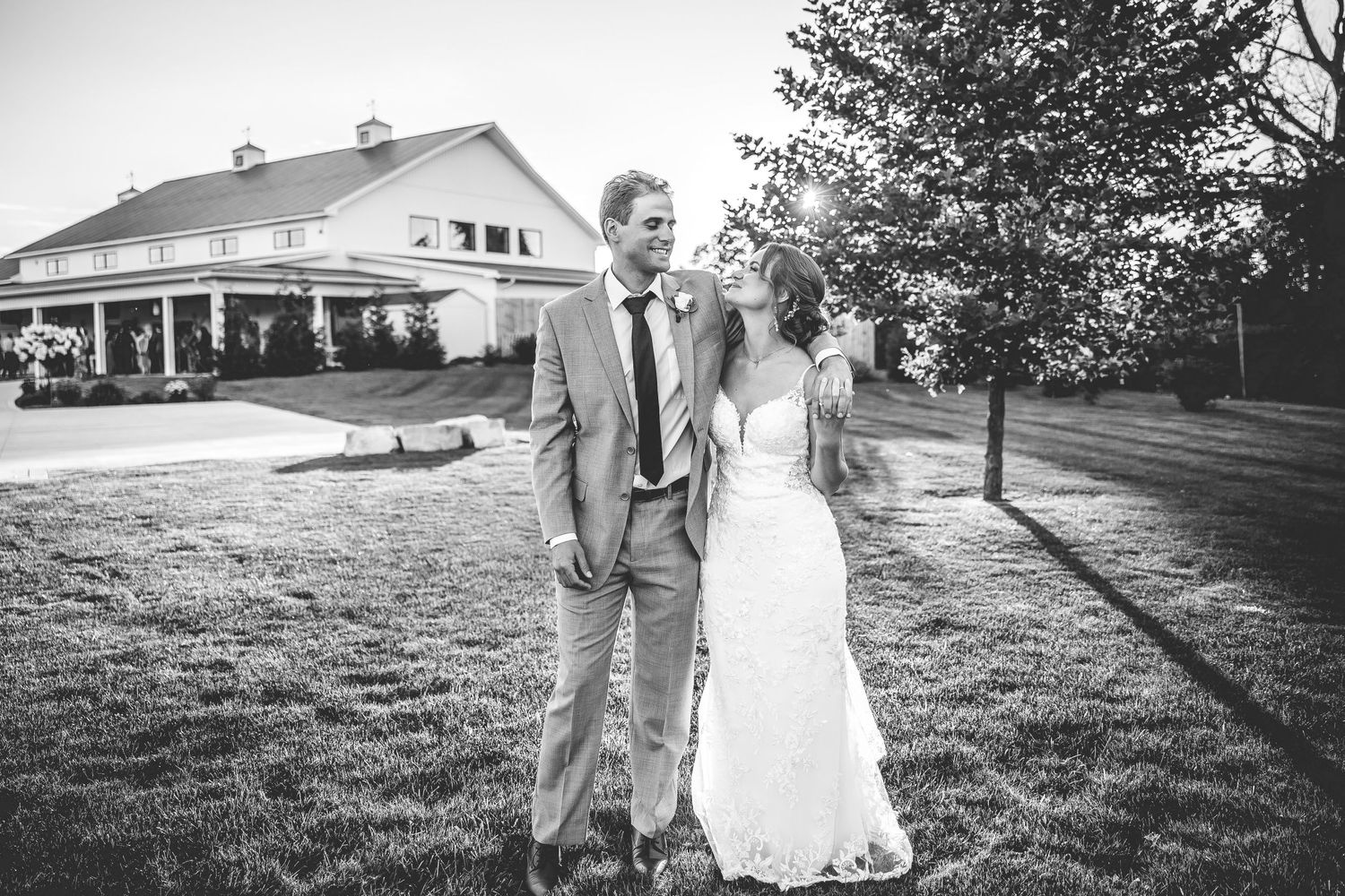 A black and white wedding photo of a couple walking on grass with a white house in the background.
