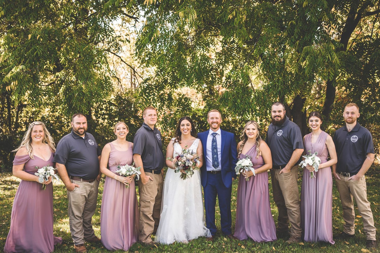 Full wedding party in mauve dresses and casual attire gathered for outdoor group portrait.