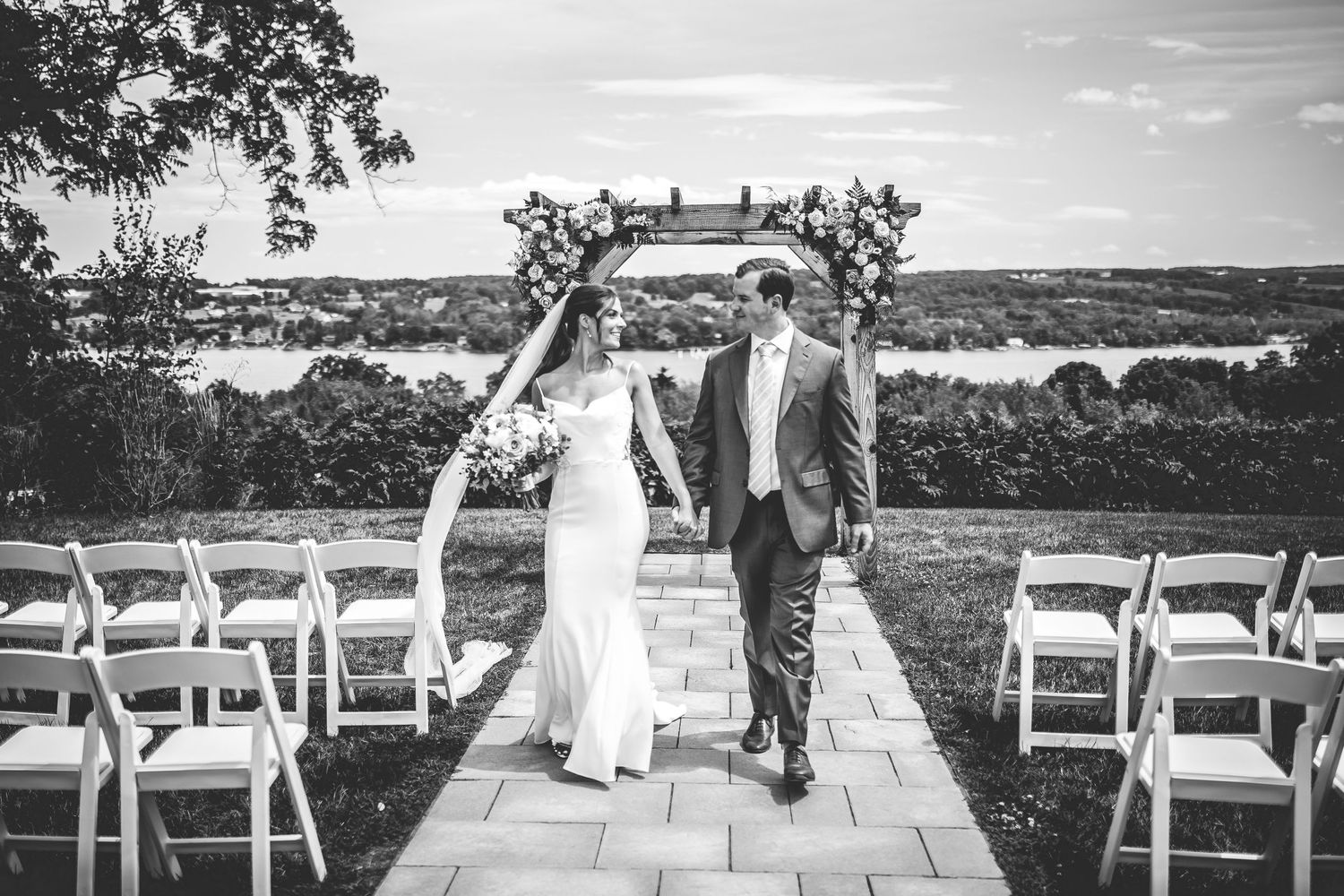 Black and white photo of an outdoor wedding ceremony setup with white chairs and wooden arch.