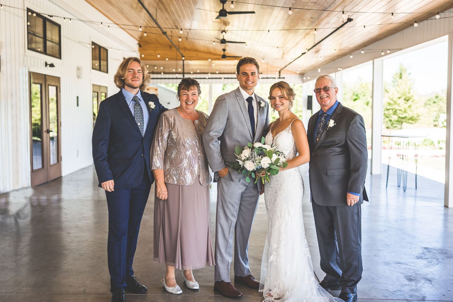A formal wedding party poses together in a long rustic venue with wooden ceiling beams.
