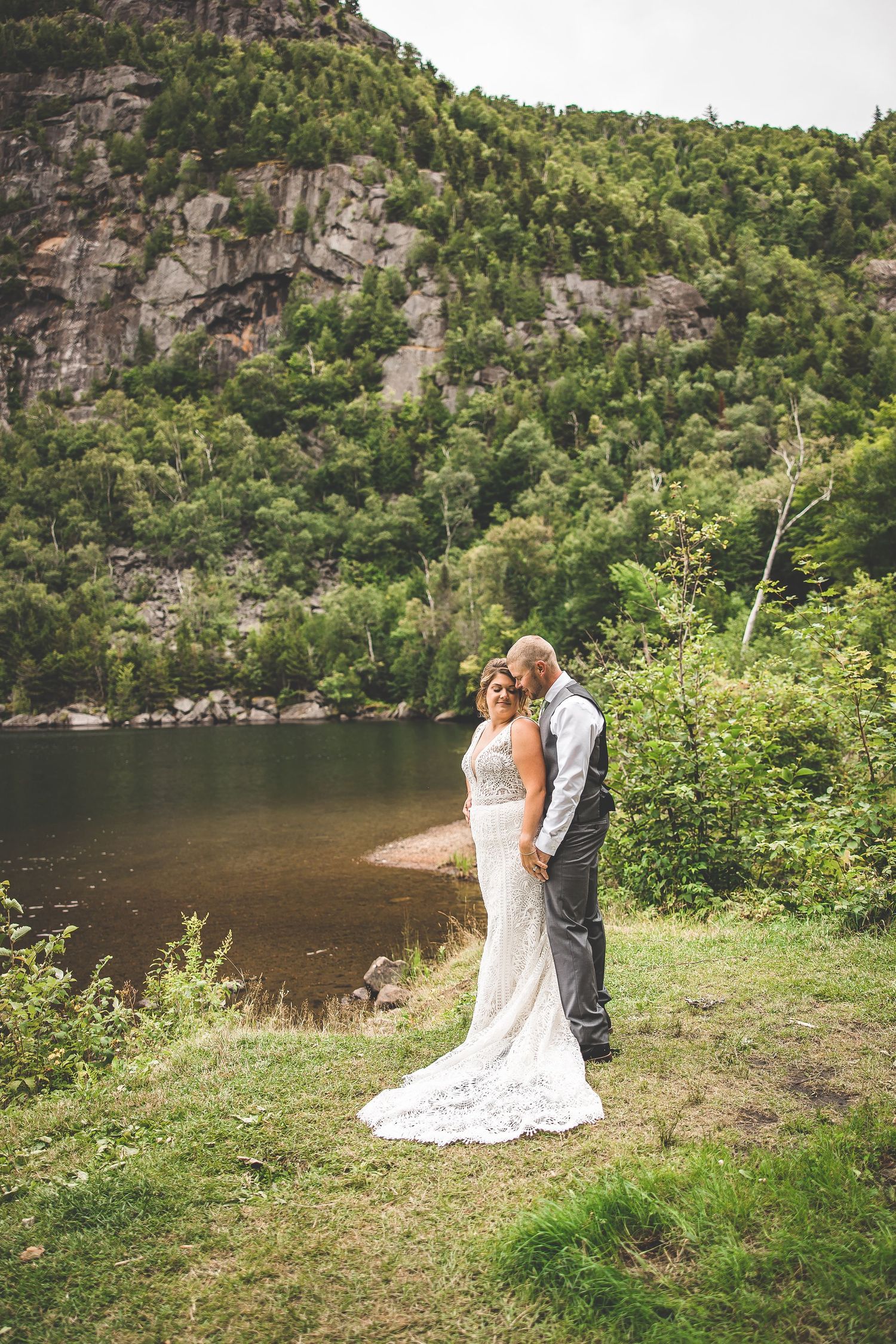 A wedding couple embraces on the shore of a mountain lake with dramatic cliffs in the background.