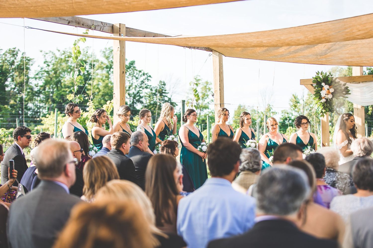 Outdoor wedding ceremony under tan canopy shades with guests standing and watching the proceedings.