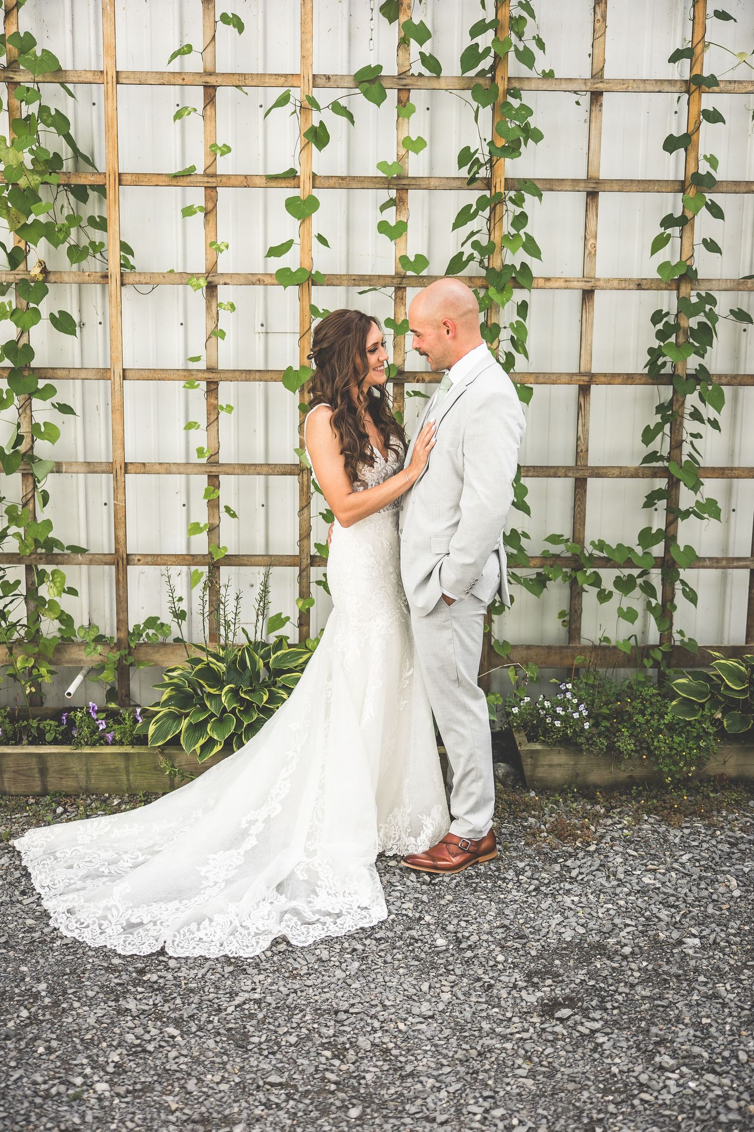 A couple poses against a white trellis wall with climbing green vines.