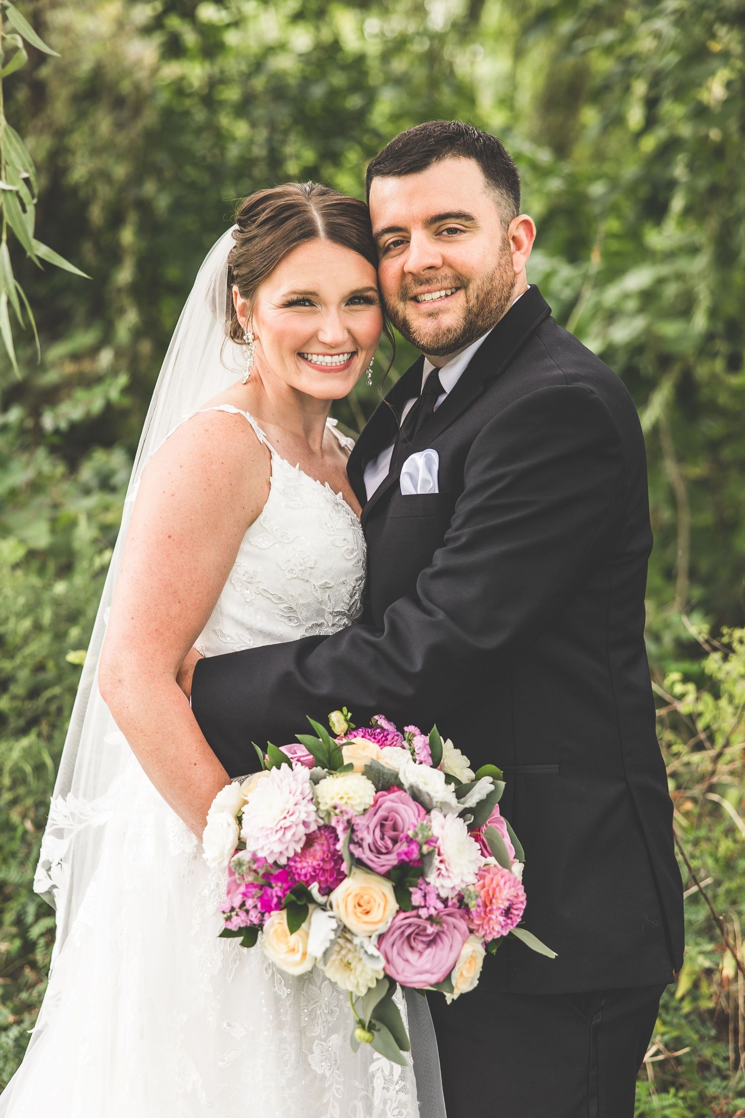 A bride in white holds a vibrant pink and purple bouquet while posing with her groom outdoors.