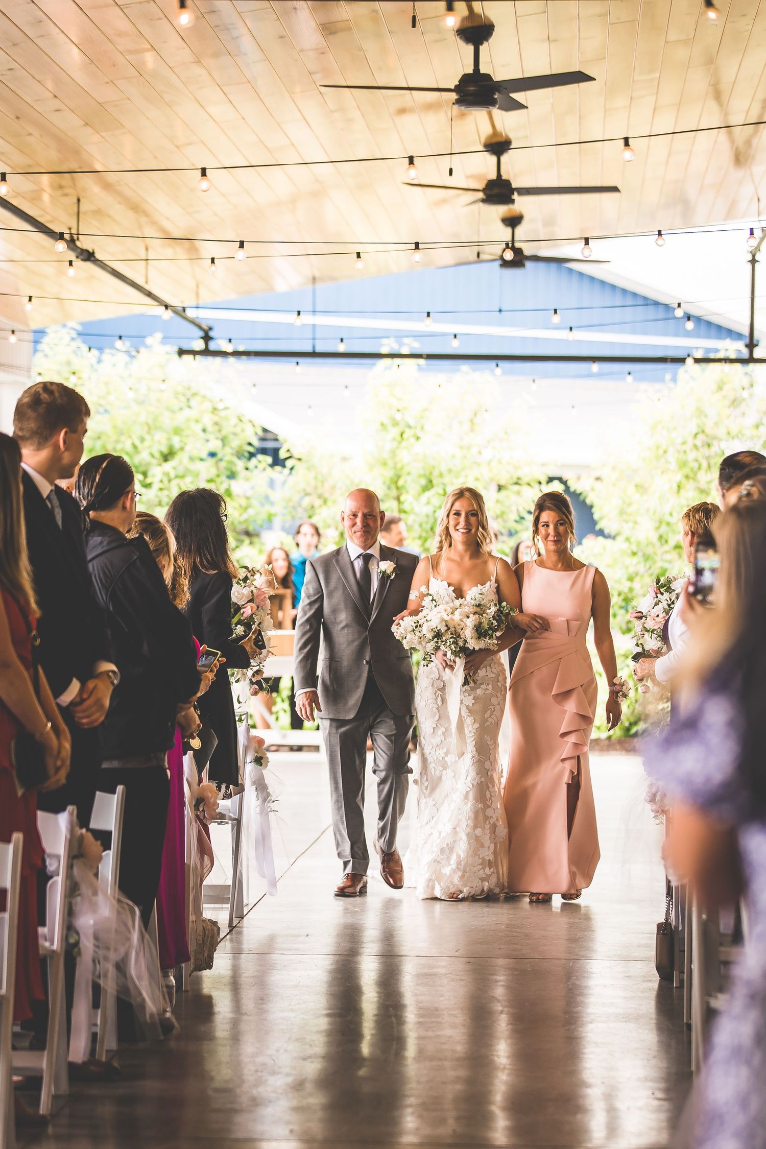 Wedding party walks down aisle in venue with string lights and wooden ceiling.