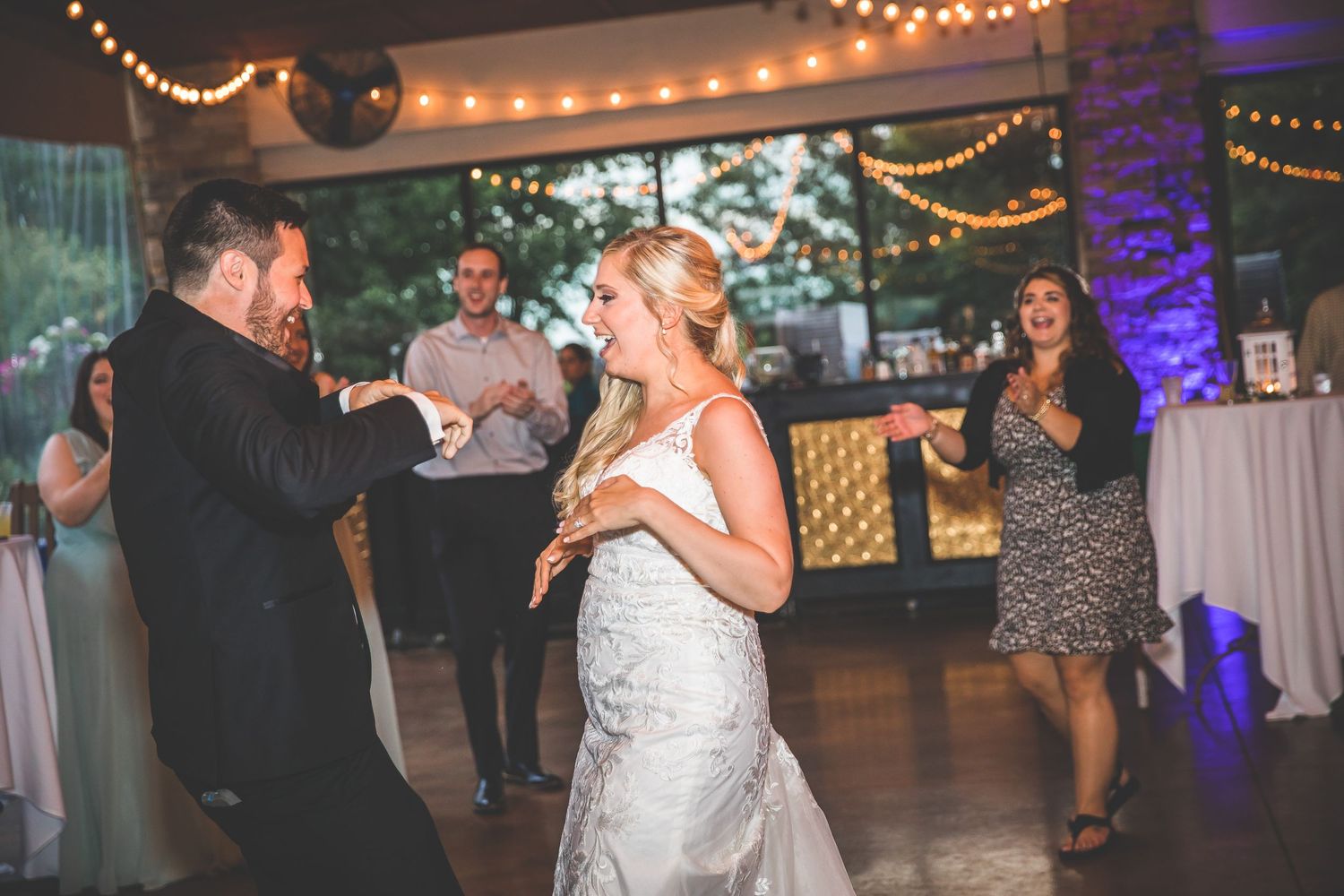 Wedding reception dance floor filled with guests under warm string lights and purple uplighting.